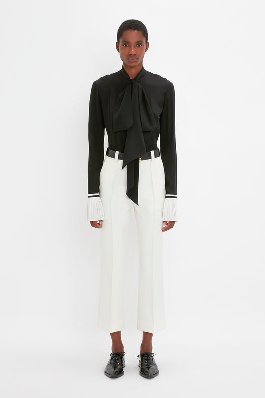 A black woman standing in a studio, wearing a black blouse with a bow and white Cropped Kick Trouser In Ivory trousers from Victoria Beckham, paired with Pointy Toe Flat Lace Up shoes.