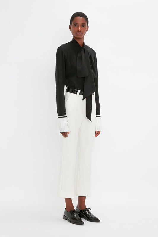 Woman standing in a studio, wearing a black blouse with bow, Victoria Beckham Cropped Kick Trouser In Ivory, and black shoes.