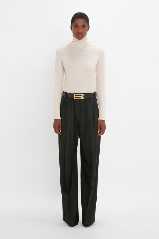 A woman wearing a Victoria Beckham cream polo neck jumper and black wide-leg trousers with a gold buckle belt, standing against a white background.