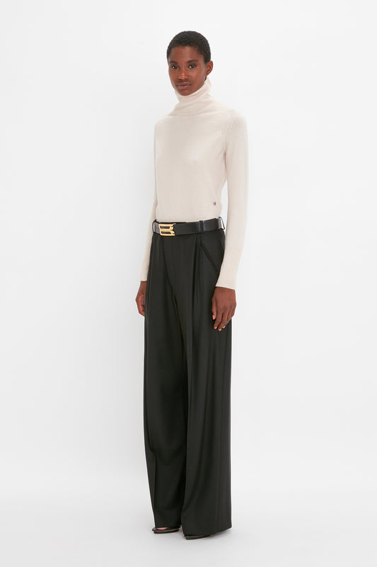 A woman wearing a Victoria Beckham cream Polo Neck Jumper In Ivory and black wide-leg trousers with a gold buckle belt, standing against a white background.