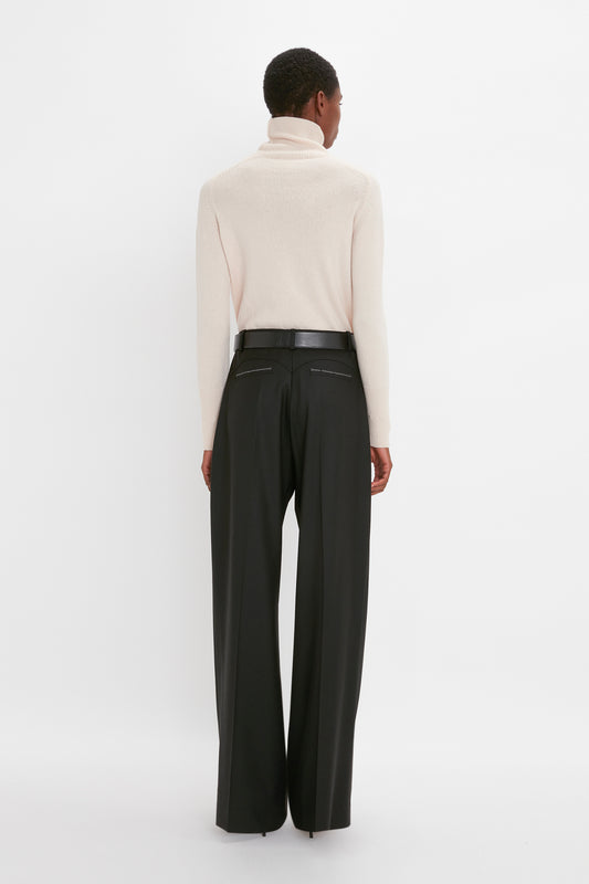 A woman stands with her back to the camera, wearing a beige Victoria Beckham polo neck jumper and black wide-leg trousers against a white background.
