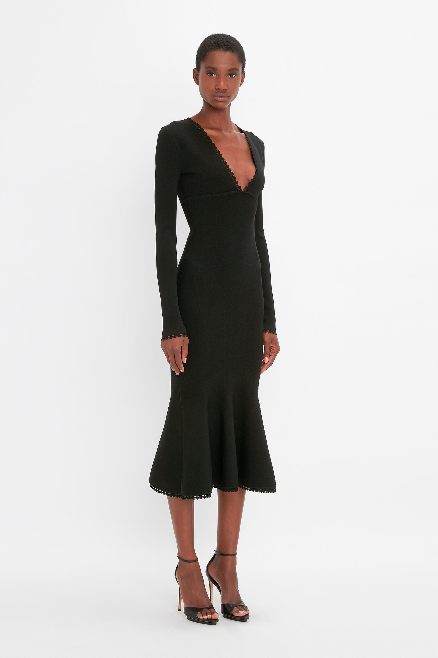 A woman is standing against a white background wearing the VB Body Long Sleeve V Neck Dress In Black by Victoria Beckham, paired with black high-heeled sandals.