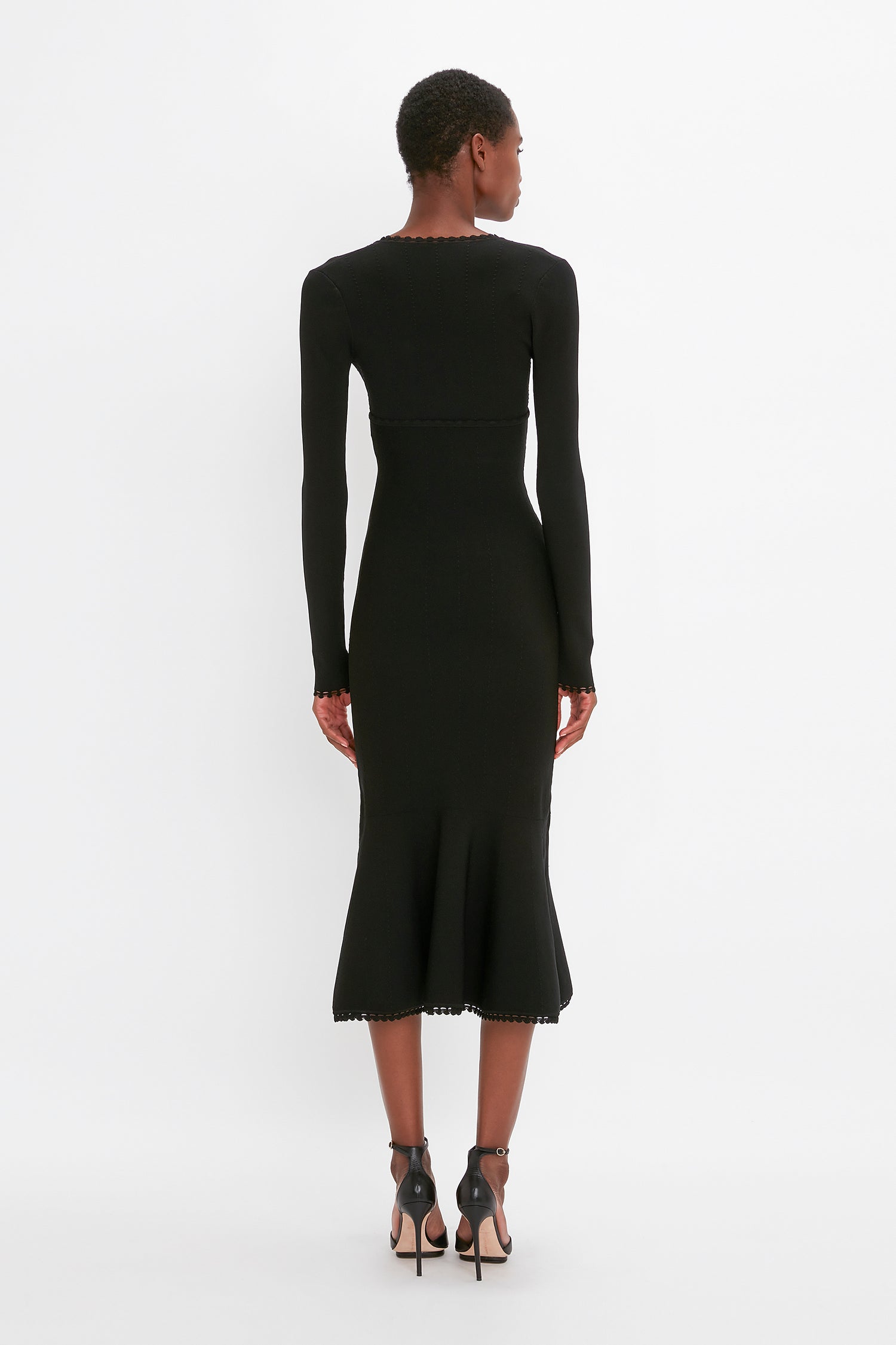 Person wearing a VB Body Long Sleeve V Neck Dress In Black by Victoria Beckham and black high heels, standing and facing away from the camera against a white background.