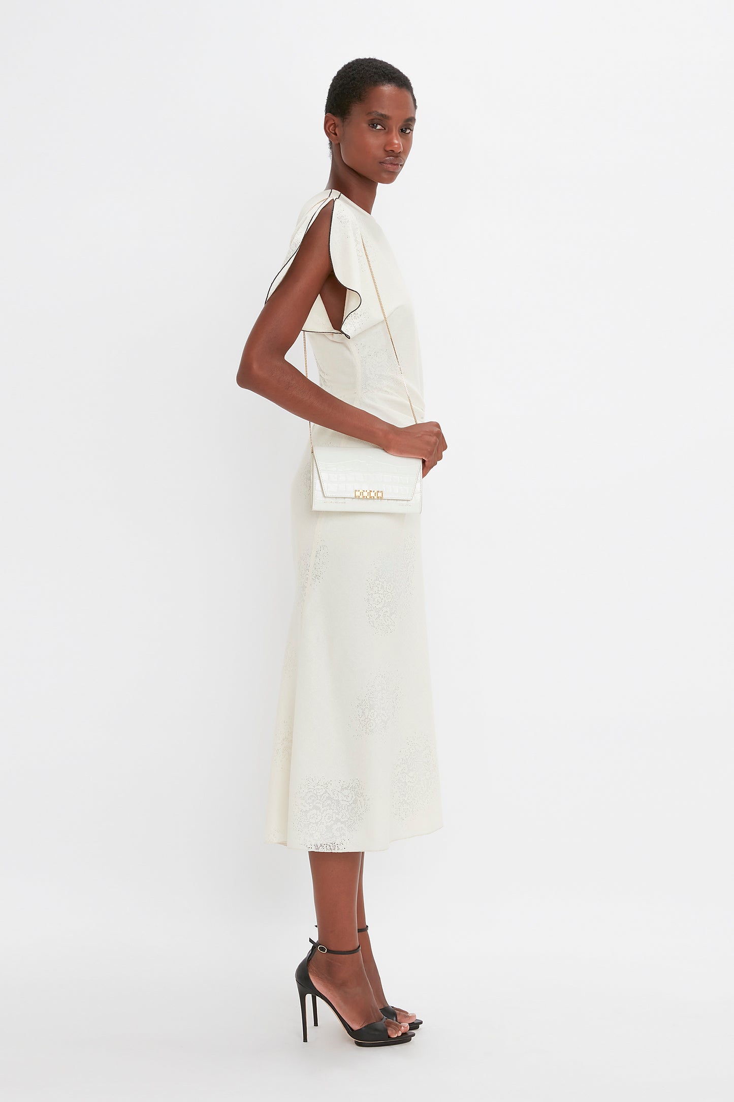 Person in a white sleeveless dress and black high heels stands facing left. They carry an Exclusive Wallet On Chain In Ivory Croc-Effect Leather by Victoria Beckham over their right shoulder and have short, close-cropped hair.