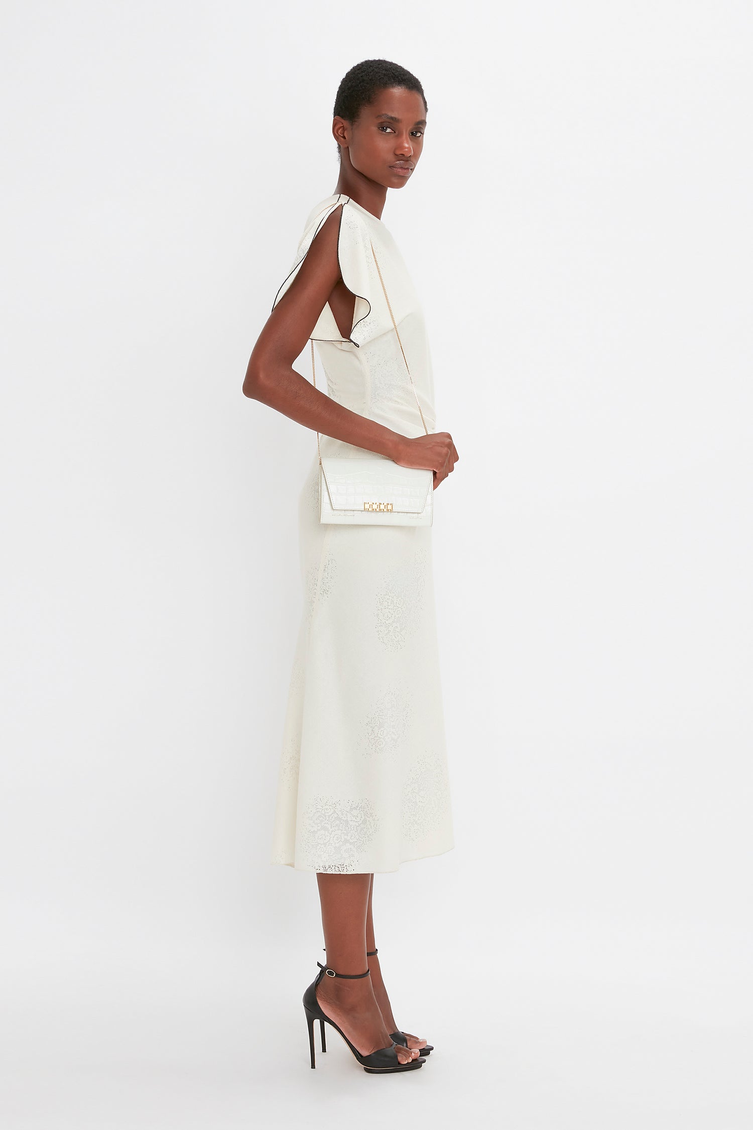 Person in a white sleeveless dress and black high heels stands facing left. They carry an Exclusive Wallet On Chain In Ivory Croc-Effect Leather by Victoria Beckham over their right shoulder and have short, close-cropped hair.