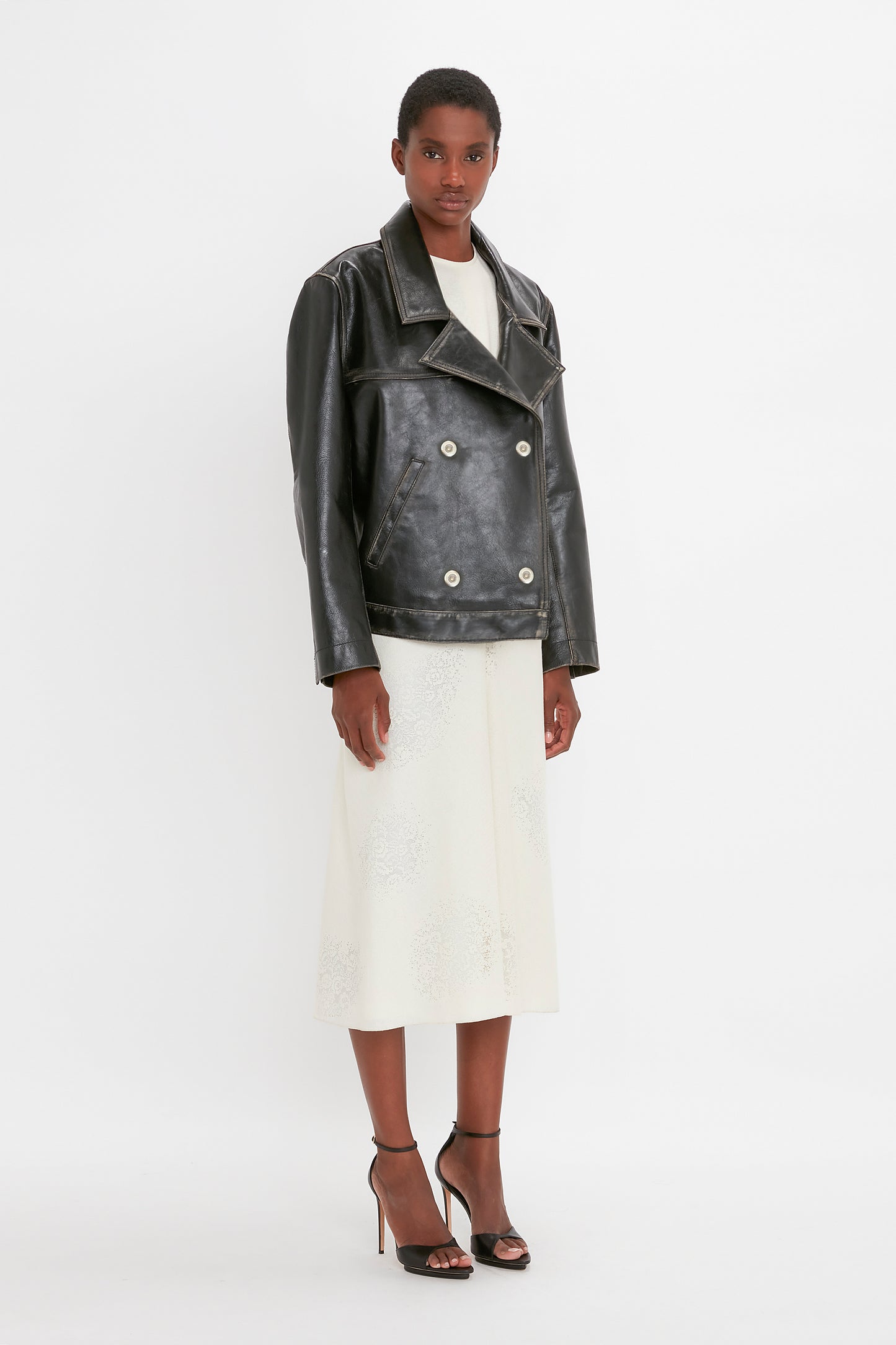 A person stands against a plain background, wearing the Victoria Beckham Oversized Leather Jacket In Black, a white knee-length skirt, and black high heels.