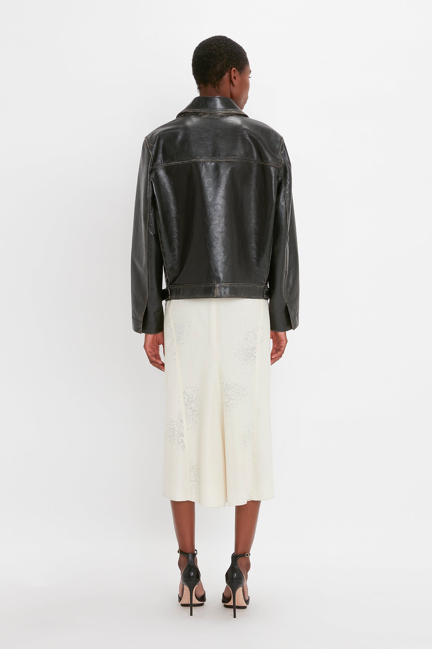 A person stands facing away, wearing an Oversized Leather Jacket In Black by Victoria Beckham, a white midi skirt, and black high-heeled sandals. The background is plain white.