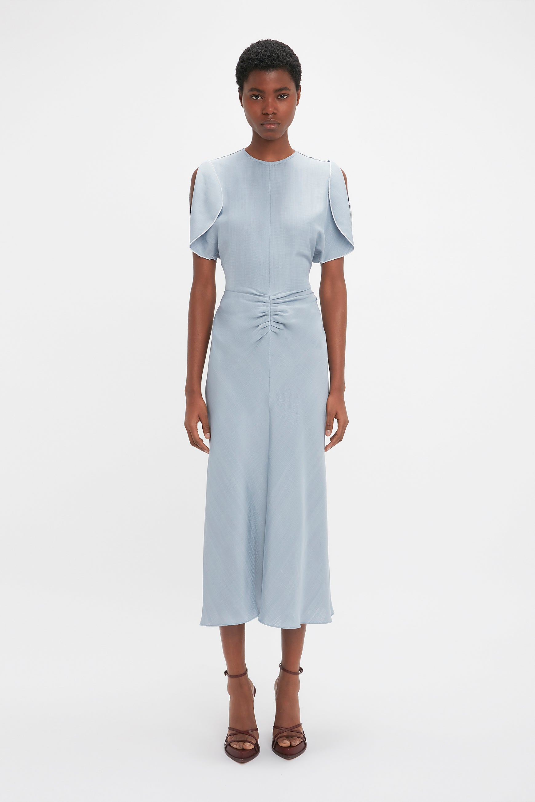 A black woman in a light blue Exclusive Gathered Waist Midi Dress In Pebble by Victoria Beckham, with cold-shoulder sleeves and standing against a white background.