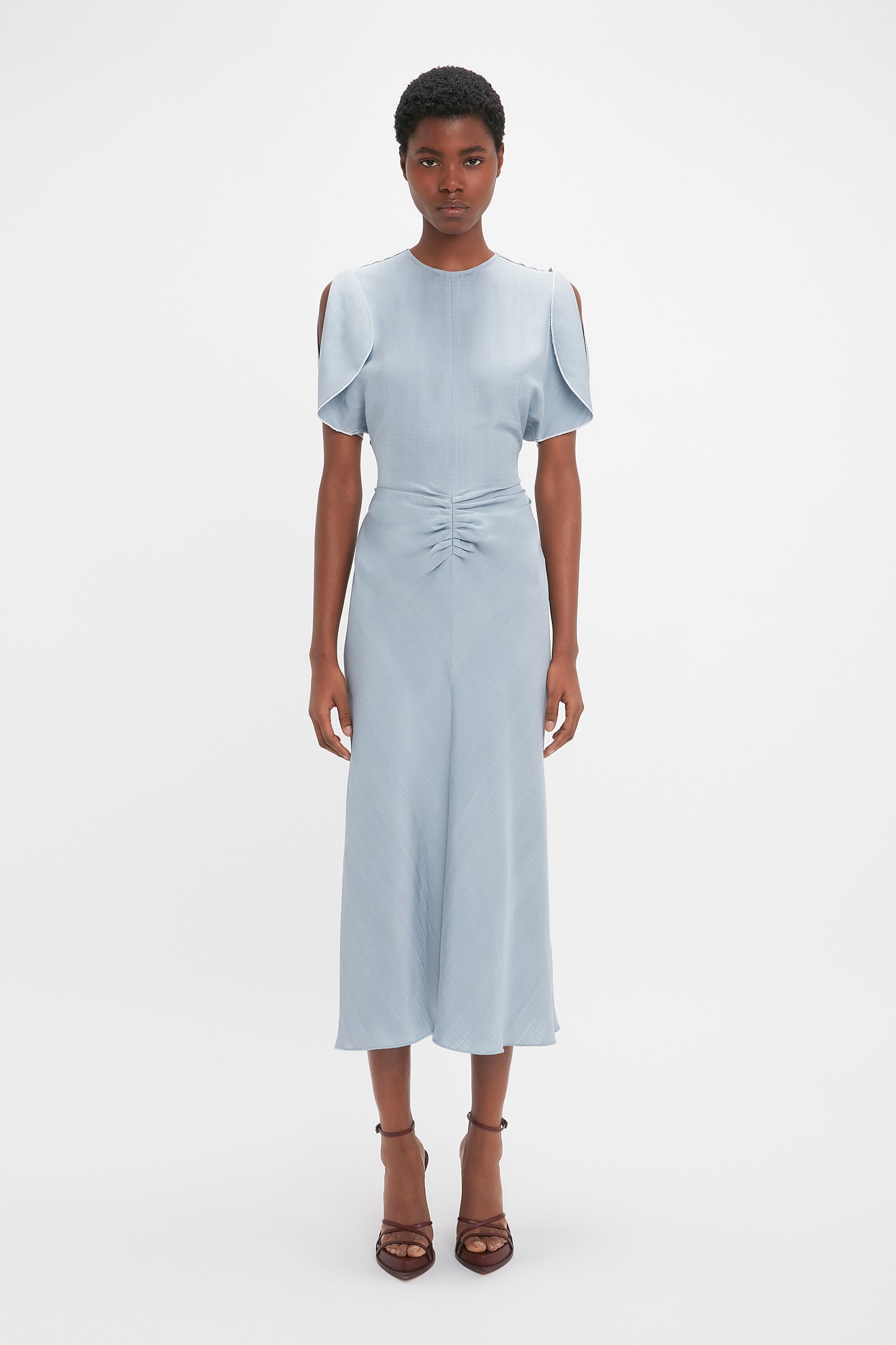 A black woman in a light blue Exclusive Gathered Waist Midi Dress In Pebble by Victoria Beckham, with cold-shoulder sleeves and standing against a white background.