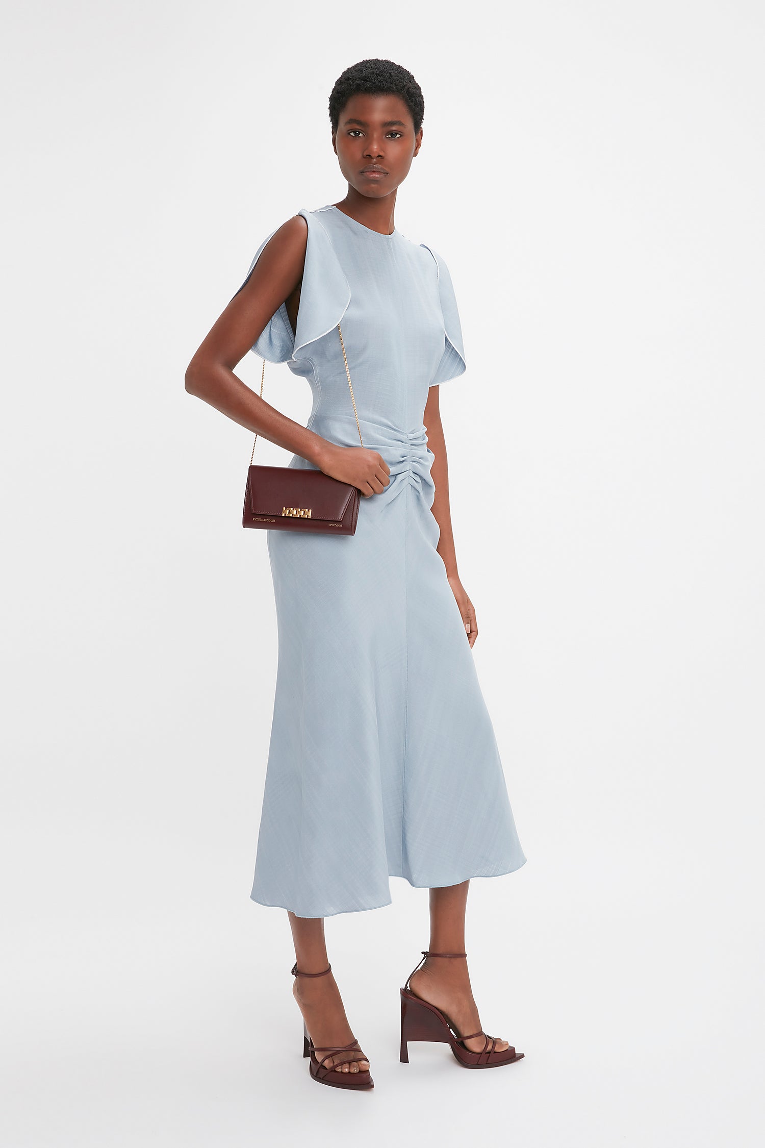 A woman stands against a plain white background wearing a light blue dress with short, ruffled, open sleeves, brown high-heeled sandals, and carrying a Victoria Beckham Wallet On Chain In Burgundy Leather.