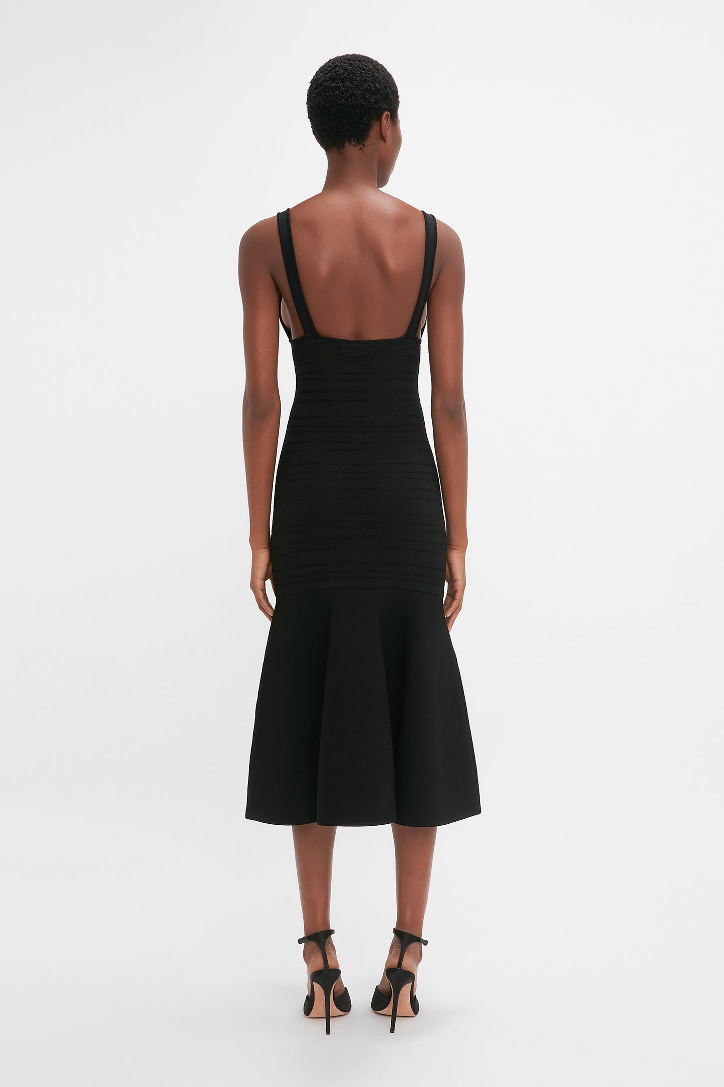 A woman seen from behind wearing a black, sleeveless fit-and-flare midi-length dress with Victoria Beckham's Frame Detail Sleeveless Dress In Black, paired with black high heels on a white background.