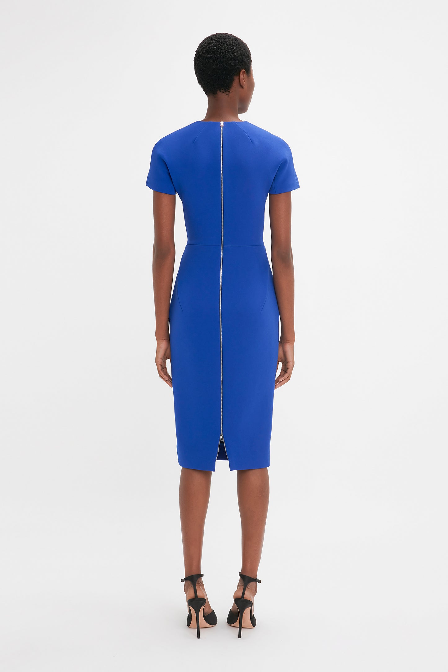 A woman from behind wearing a Victoria Beckham Fitted T-Shirt Dress in Palace Blue and black high heels, standing against a white background.