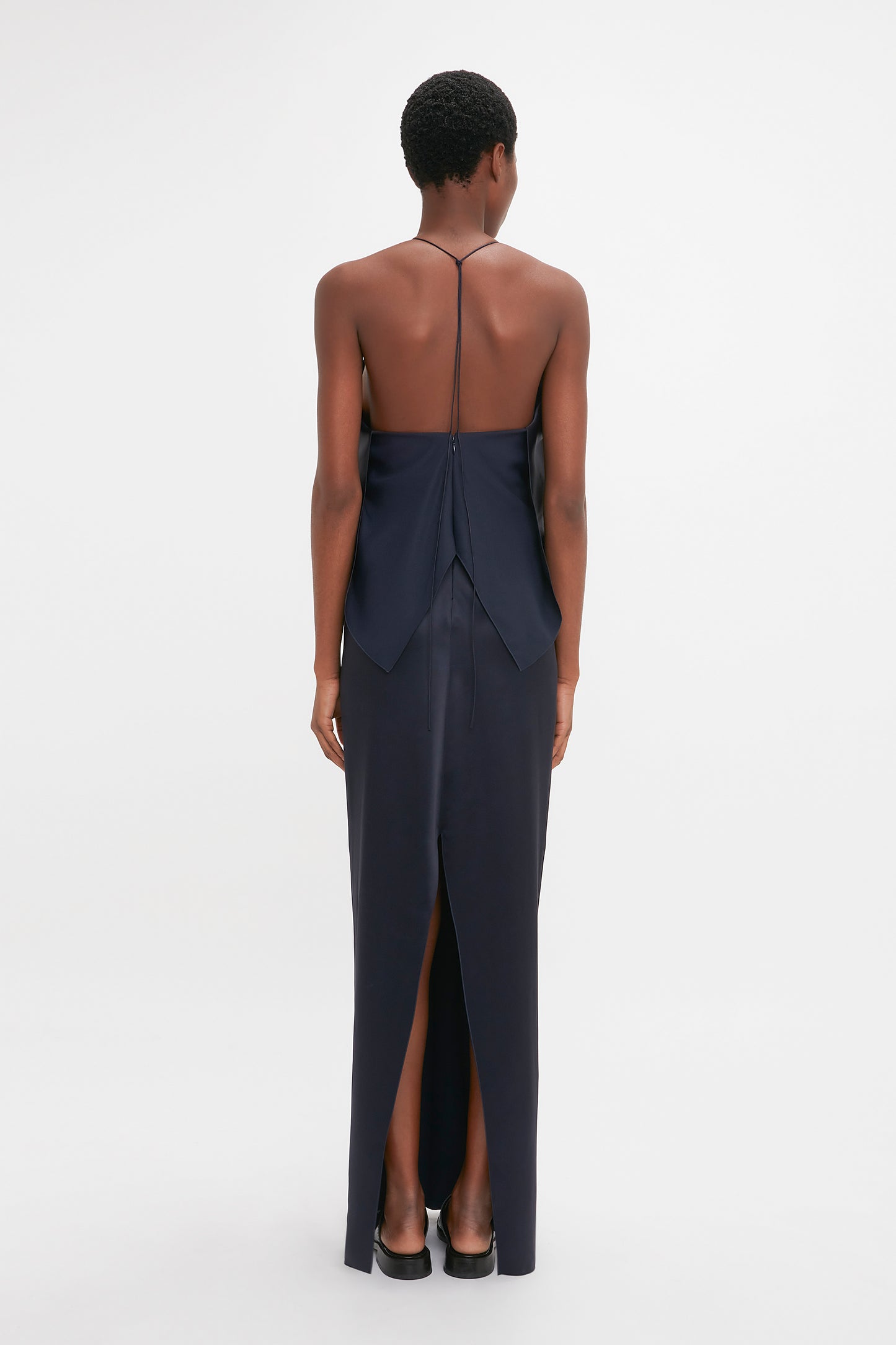 A woman viewed from behind wearing a Victoria Beckham navy backless evening gown with a delicate tie, standing against a white background.