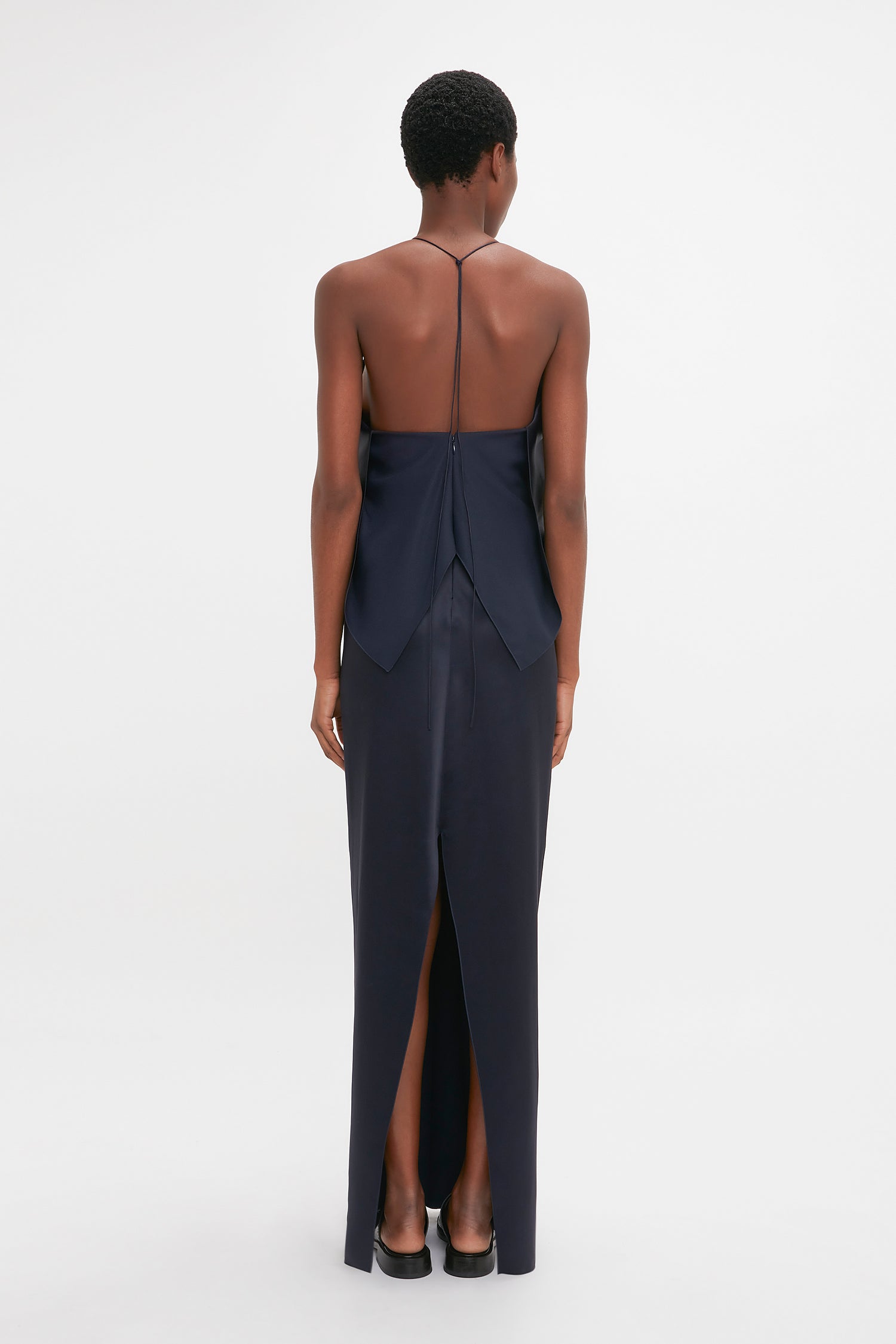 A woman viewed from behind wearing a Victoria Beckham navy backless evening gown with a delicate tie, standing against a white background.