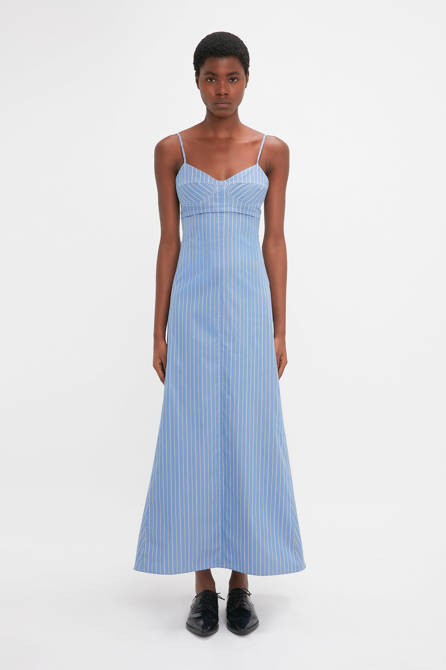 A person is wearing a Cami Fit And Flare Midi In Steel Blue by Victoria Beckham with thin shoulder straps and Flat Lace Up Mules, standing against a plain white background.