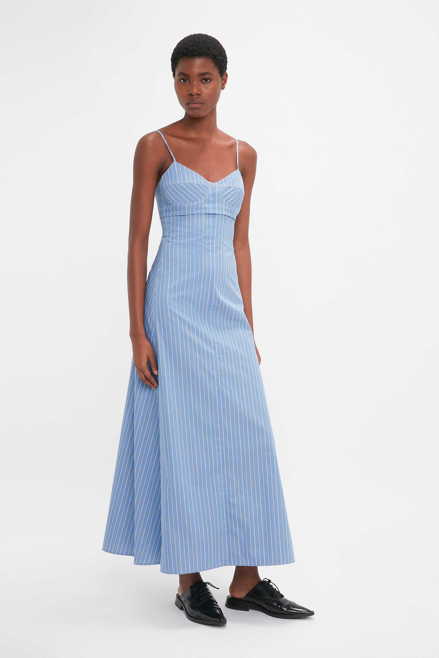 Person wearing a sleeveless, light blue striped cotton sundress with vertical white stripes and black shoes, standing against a plain white background. The dress is the Cami Fit And Flare Midi In Steel Blue by Victoria Beckham.