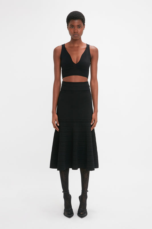 A young woman standing in a studio setting, wearing a black Victoria Beckham Frame Detail Sleeveless Top with a cropped hemline, a pleated midi skirt, patterned tights, and black boots, gazing at