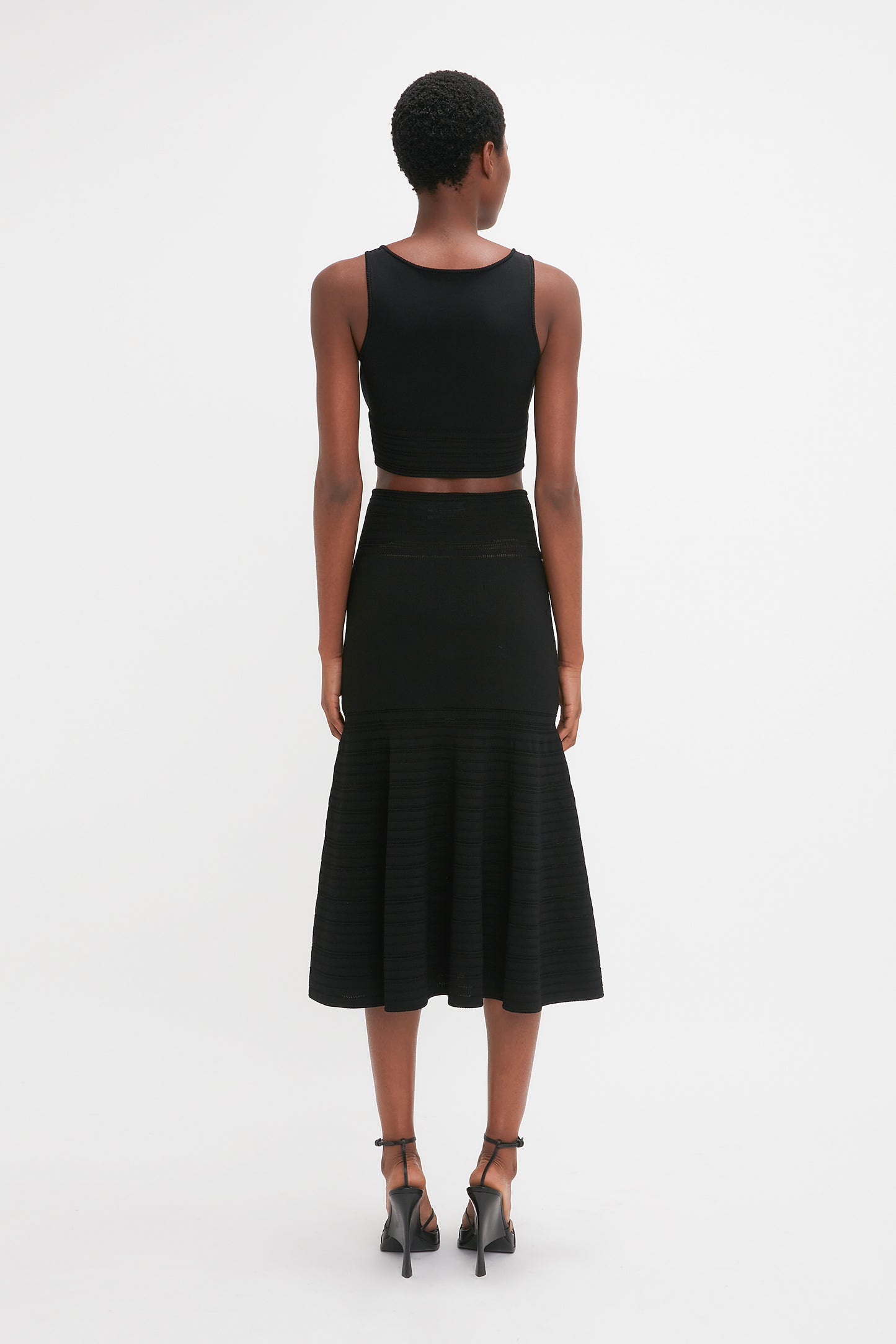 A woman wearing a black sleeveless crop top and a waist-defining Victoria Beckham Fit And Flare Midi Skirt In Black with contrasting stitching stands facing away from the camera against a plain white background.
