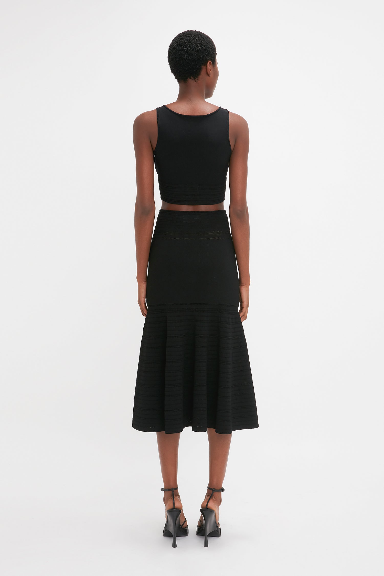 A woman wearing a black sleeveless crop top and a waist-defining Victoria Beckham Fit And Flare Midi Skirt In Black with contrasting stitching stands facing away from the camera against a plain white background.