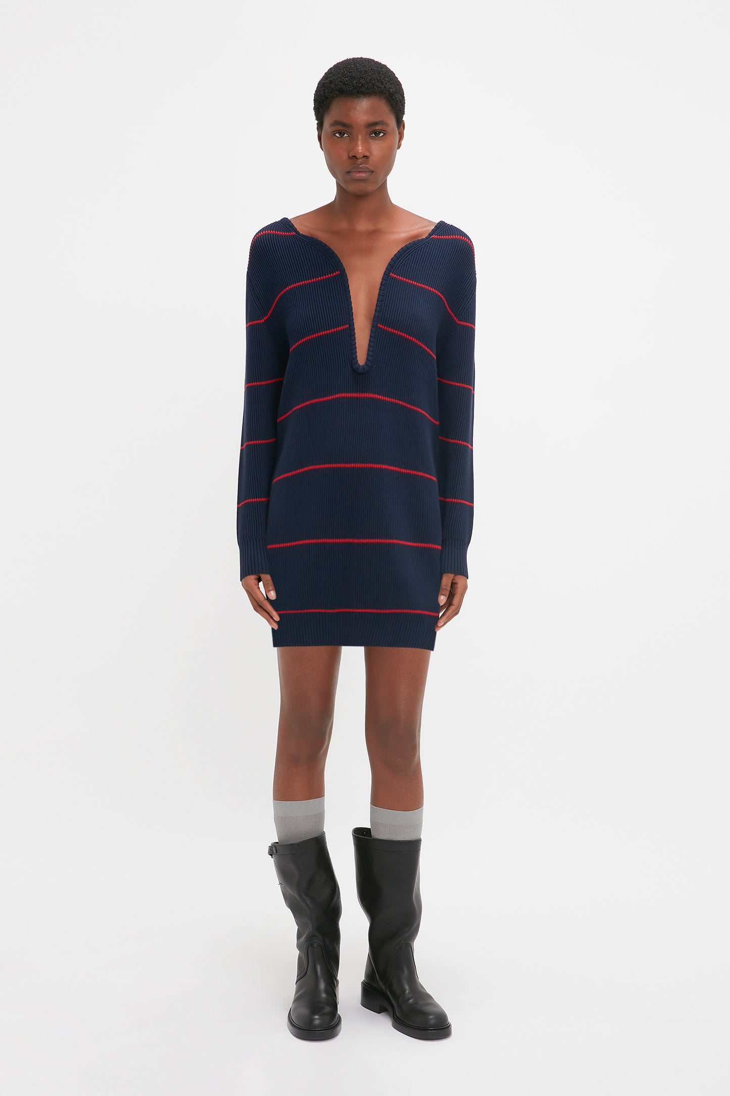 A person is wearing a Victoria Beckham Frame Detail Jumper Dress In Navy-Red with a deep V-neckline, black knee-high boots, and grey socks.