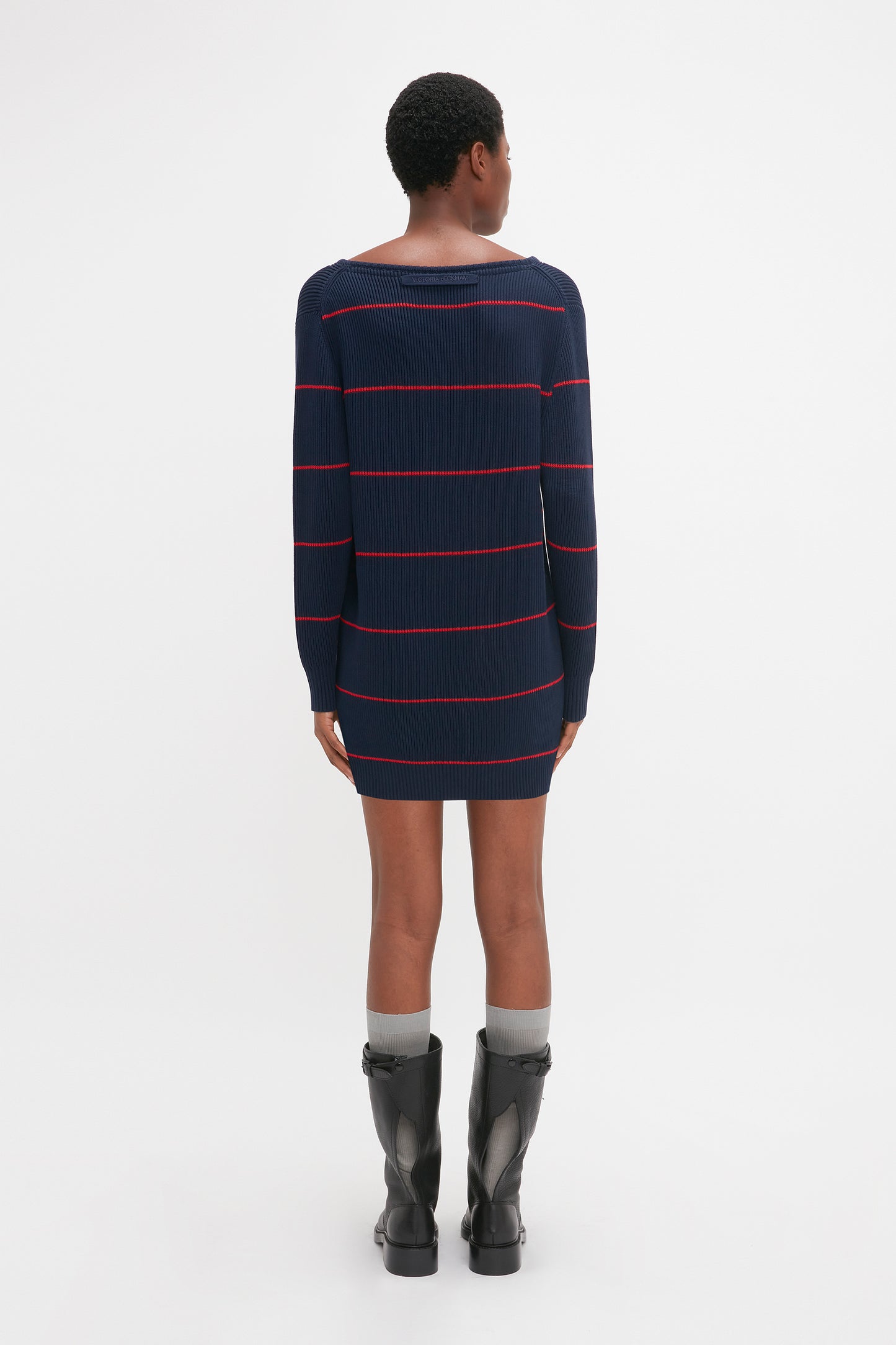 Person in a dark, long-sleeve, Frame Detail Jumper Dress In Navy-Red by Victoria Beckham stands facing away with hands by sides, wearing gray socks and black boots.