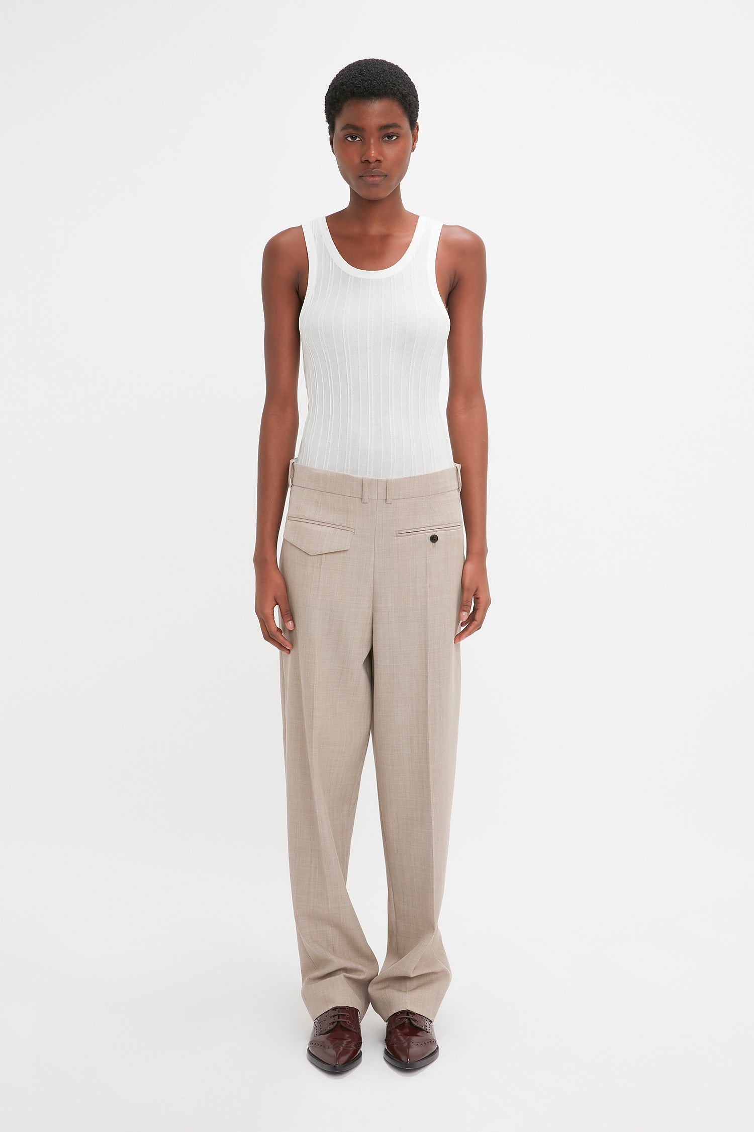 A modern woman stands against a plain background wearing a white tank top, Victoria Beckham Reverse Front Trouser In Sesame, and brown shoes, looking directly at the camera.