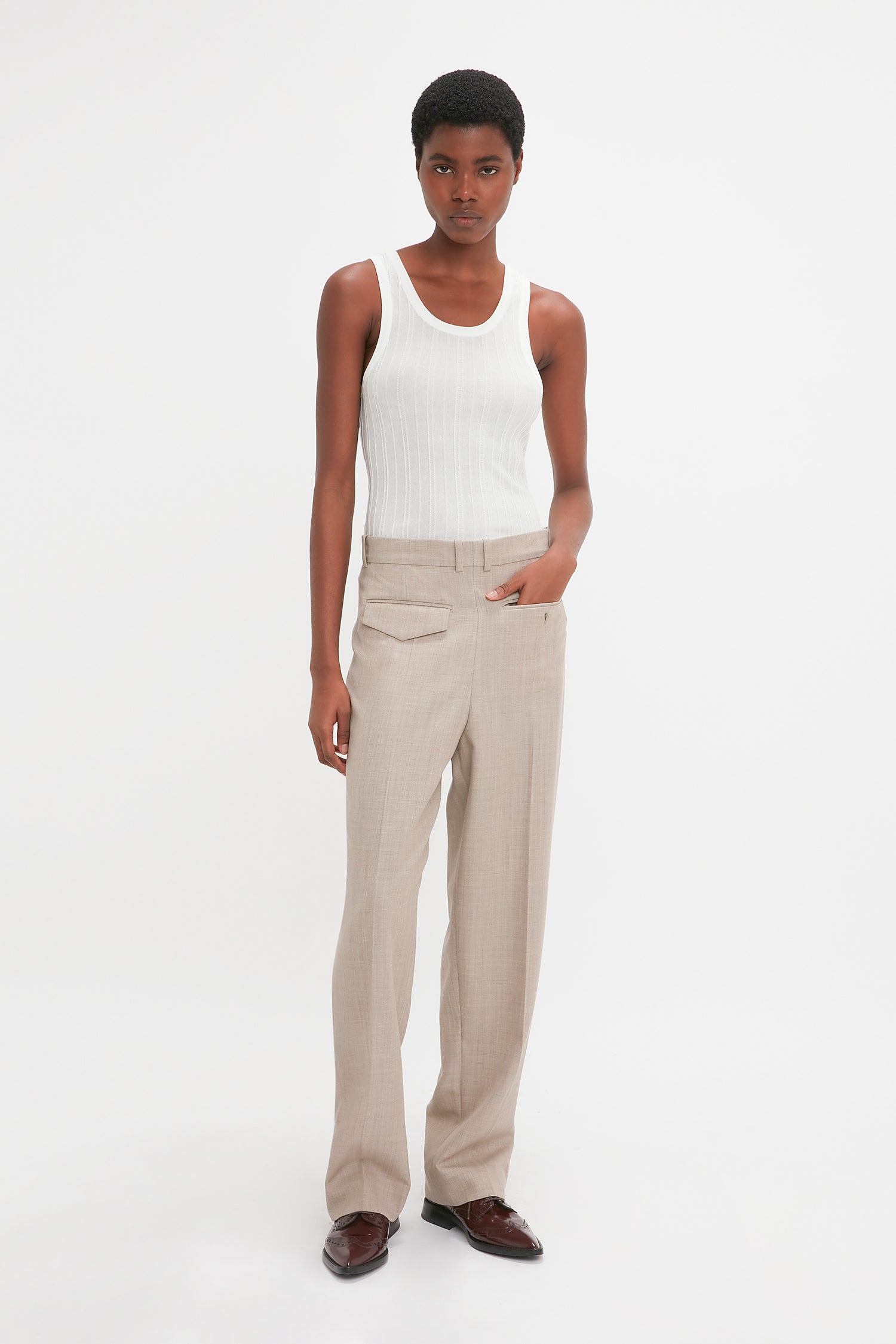 A modern woman stands against a white background, wearing a white sleeveless top, Victoria Beckham's Reverse Front Trouser In Sesame, and brown shoes. One hand is in her pocket while the other arm is relaxed at her side.