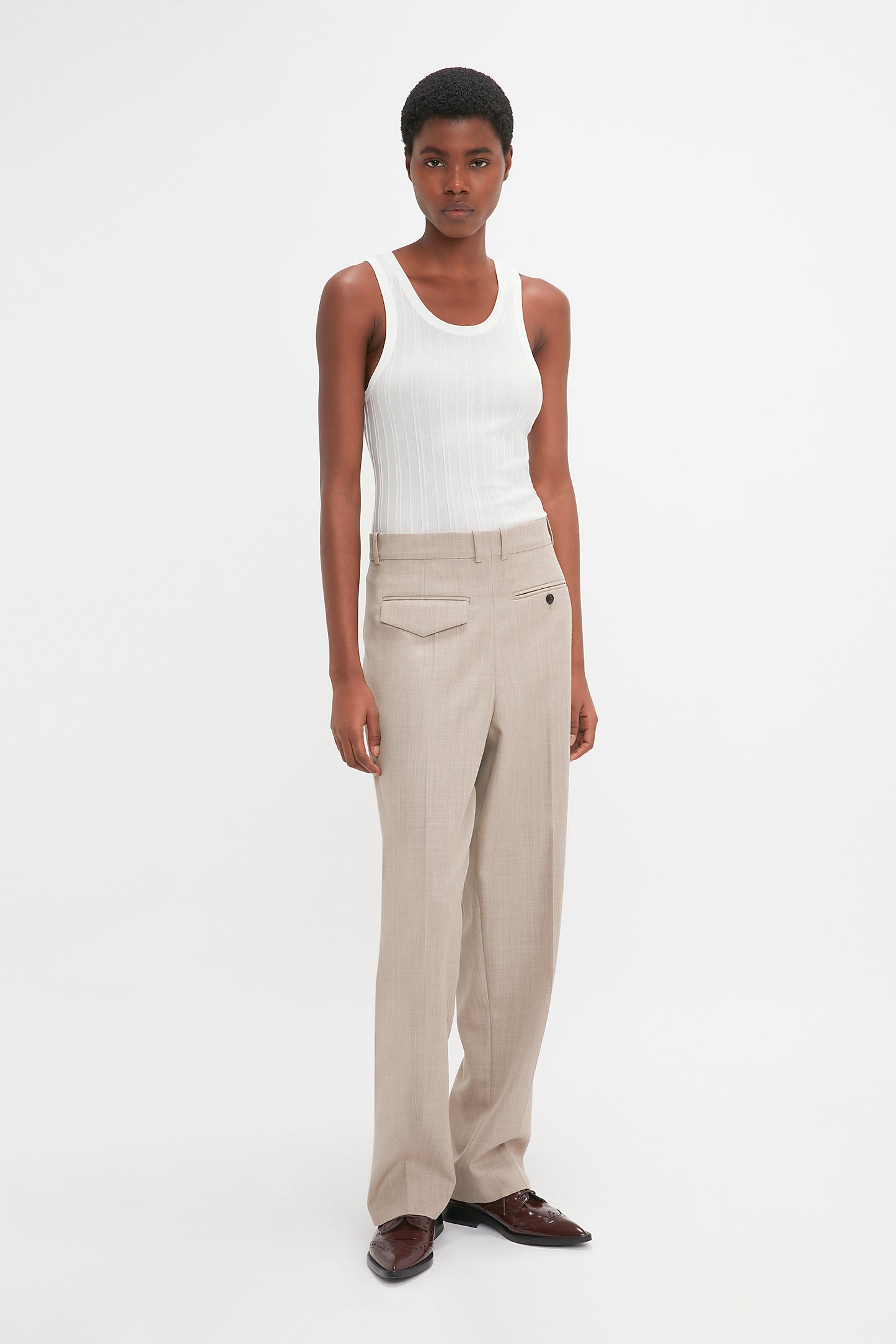 A person standing against a white background, wearing a Victoria Beckham Fine Knit Vertical Stripe Tank In White, beige trousers, and brown shoes.