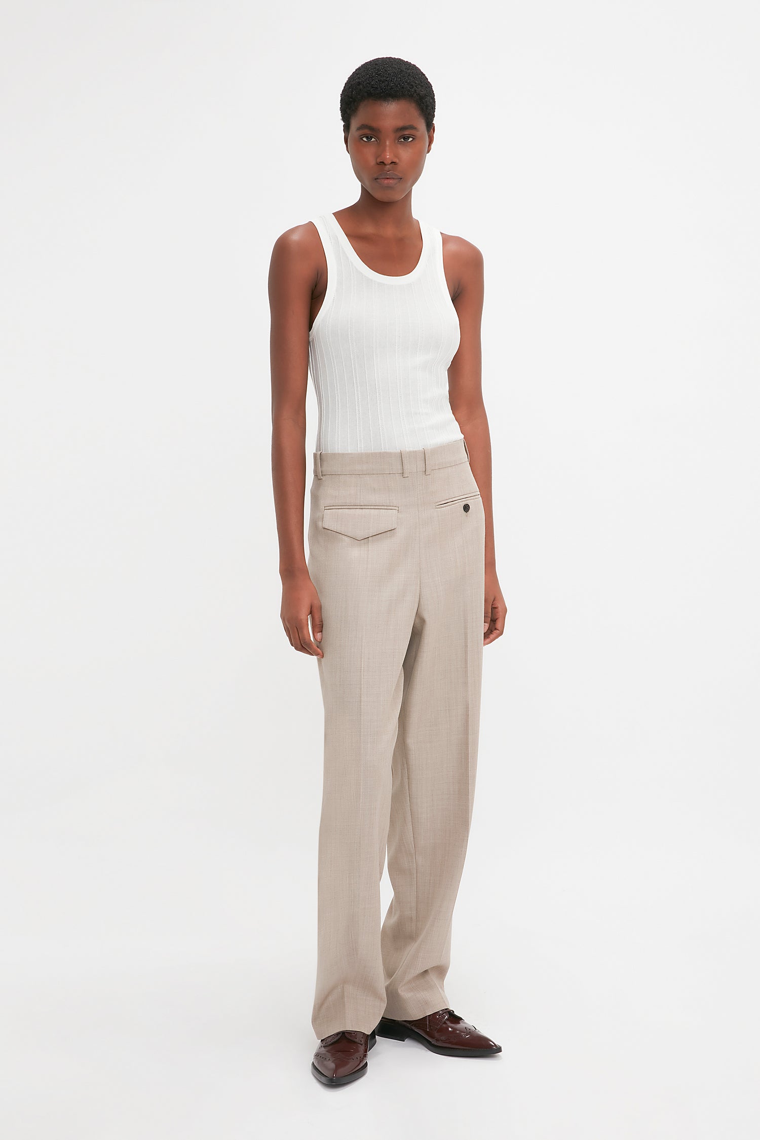 A person standing against a white background, wearing a Victoria Beckham Fine Knit Vertical Stripe Tank In White, beige trousers, and brown shoes.