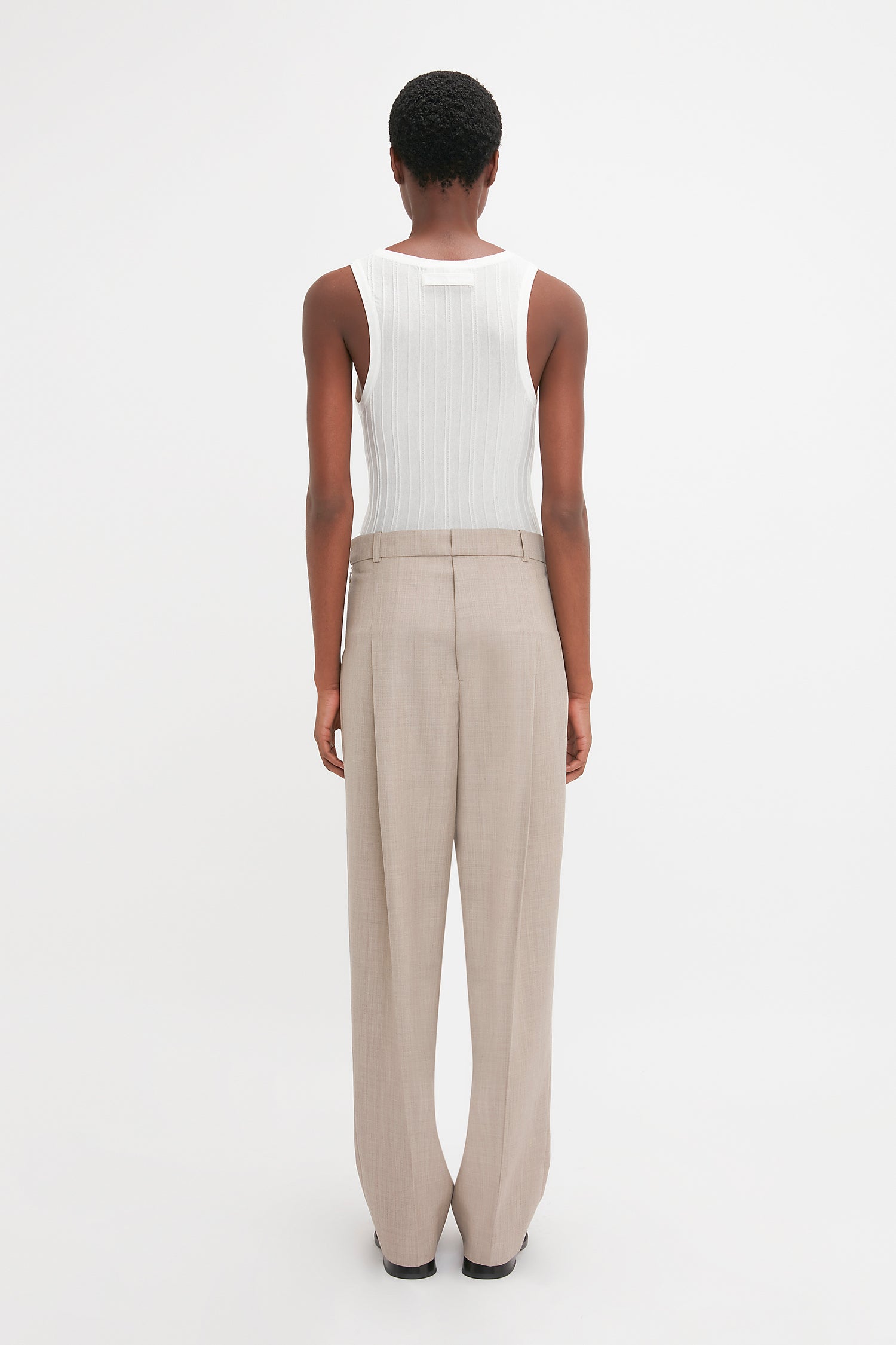 A person with short hair, wearing a Victoria Beckham Fine Knit Vertical Stripe Tank In White and beige pants, is standing and facing away from the camera against a plain white background.