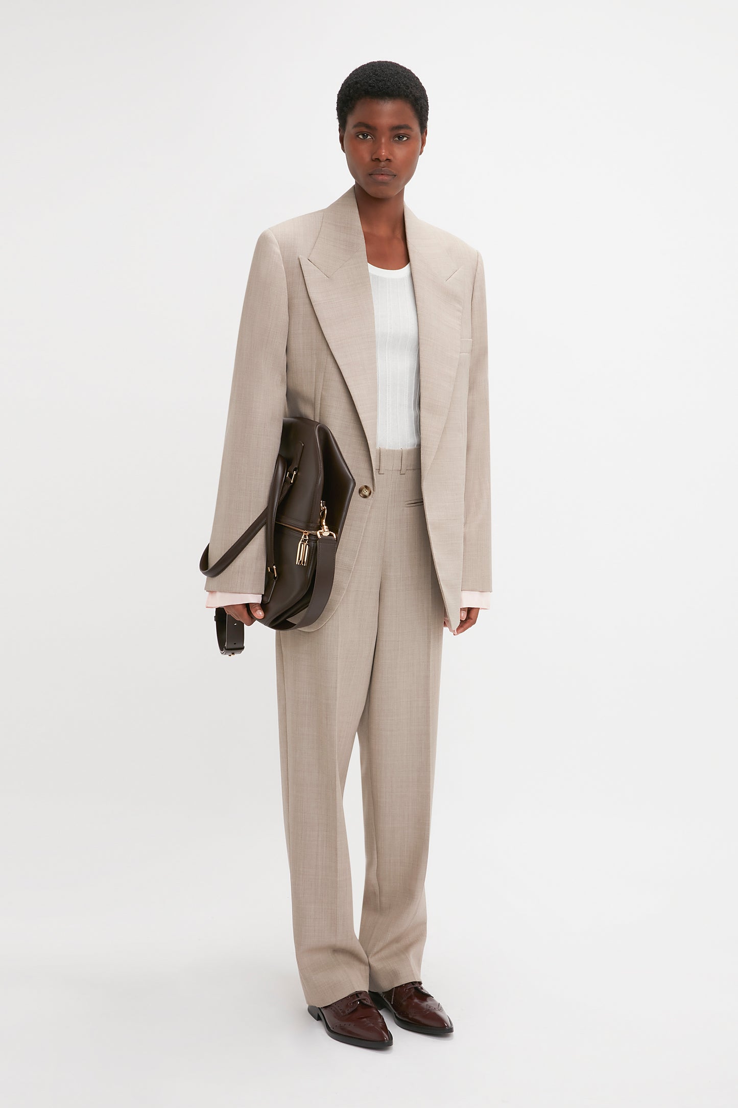 Person standing in a beige suit with a white shirt underneath, holding a dark brown briefcase, and wearing brown shoes. The Victoria Beckham Darted Sleeve Tailored Jacket In Sesame showcases contemporary detailing that enhances the overall sharp look.