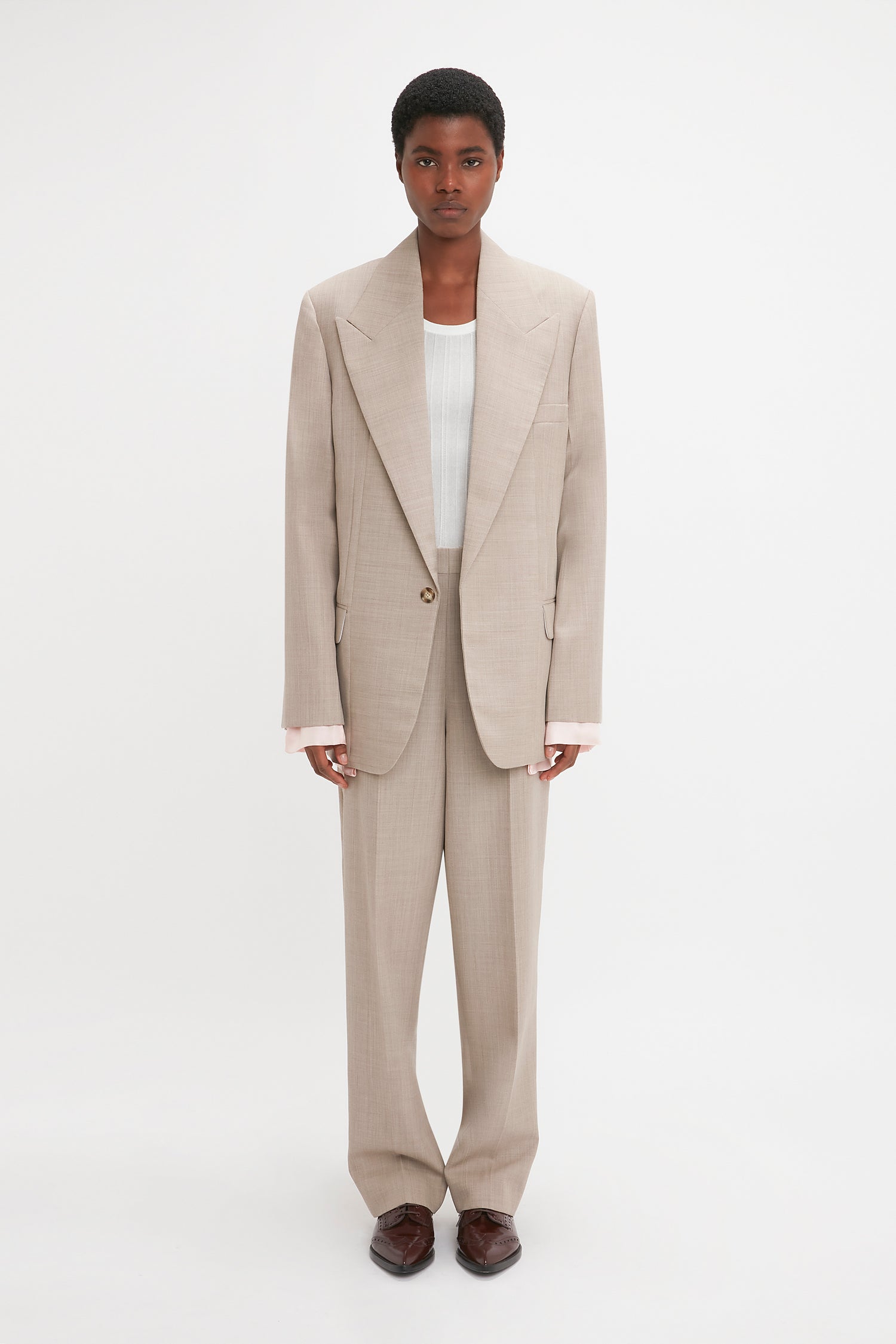 Person standing against a plain background, wearing a Darted Sleeve Tailored Jacket In Sesame by Victoria Beckham over a white top and brown shoes, showcasing contemporary detailing.