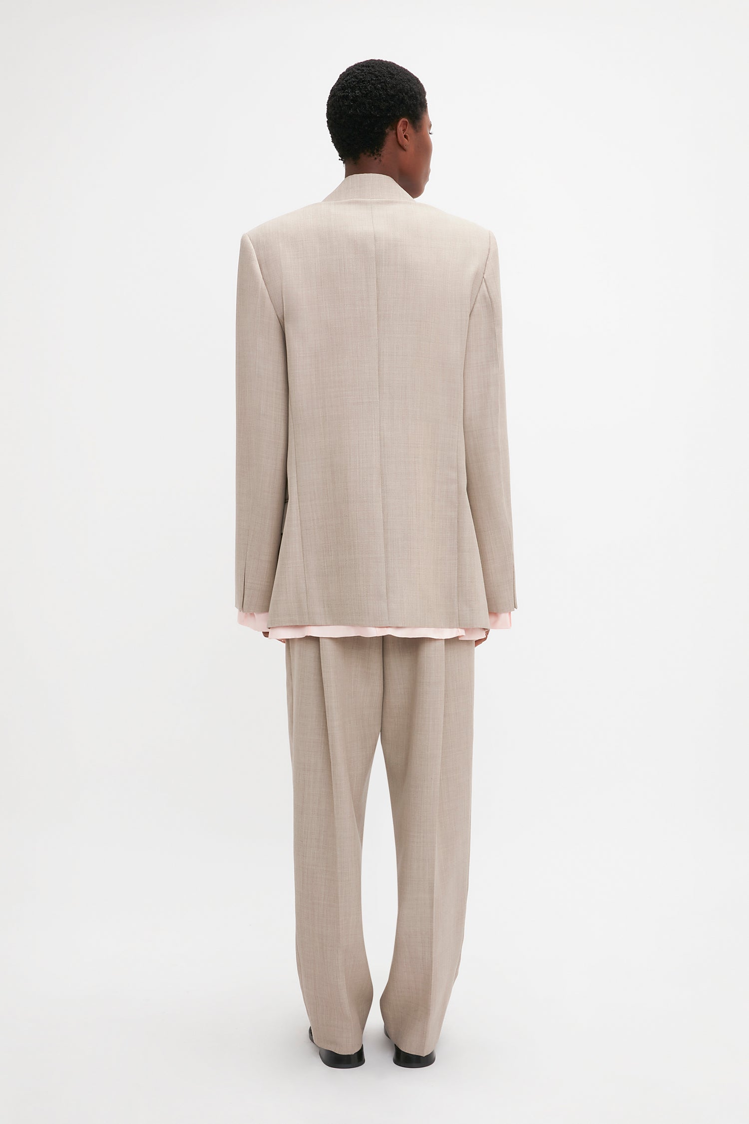 A person stands facing away from the camera, wearing a beige Darted Sleeve Tailored Jacket In Sesame from Victoria Beckham, paired with slightly loose pants and a light pink shirt beneath the blazer. The background is plain white.