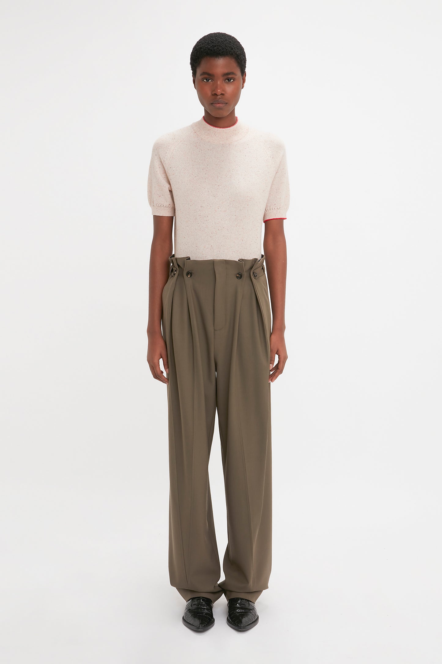 A person stands against a plain white background, wearing a Victoria Beckham Short Sleeve Top In Nougat with delicate pointelle lace stitching and high-waisted, loose-fitting khaki trousers paired with black shoes.