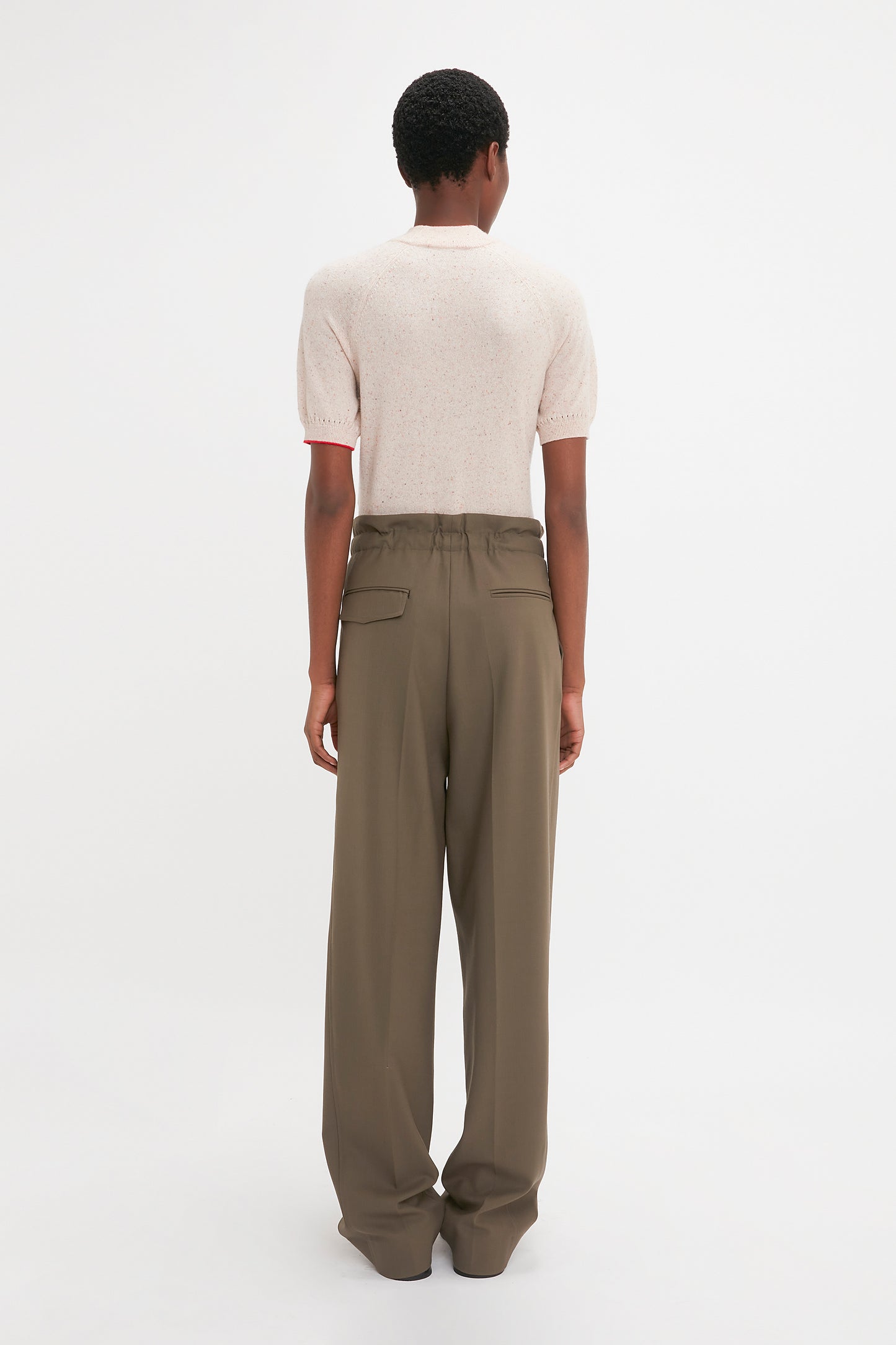 A person with short hair is standing facing away from the camera, wearing a Victoria Beckham Short Sleeve Top In Nougat and high-waisted, wide-leg olive green pants. The subtle detailing of pointelle lace stitch on the sleeves adds a touch of elegance to the outfit.