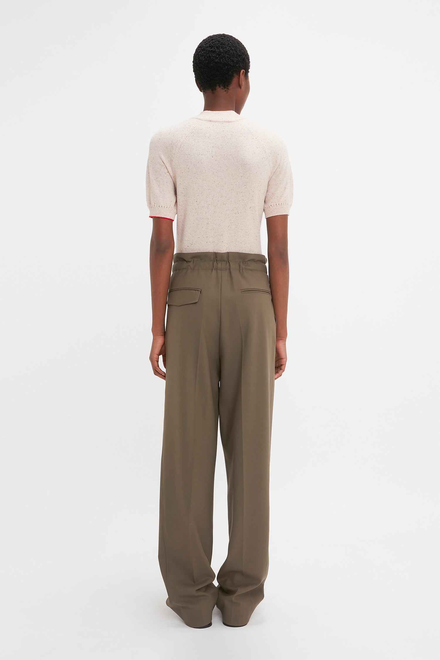 A person with short hair is standing facing away from the camera, wearing a Victoria Beckham Short Sleeve Top In Nougat and high-waisted, wide-leg olive green pants. The subtle detailing of pointelle lace stitch on the sleeves adds a touch of elegance to the outfit.