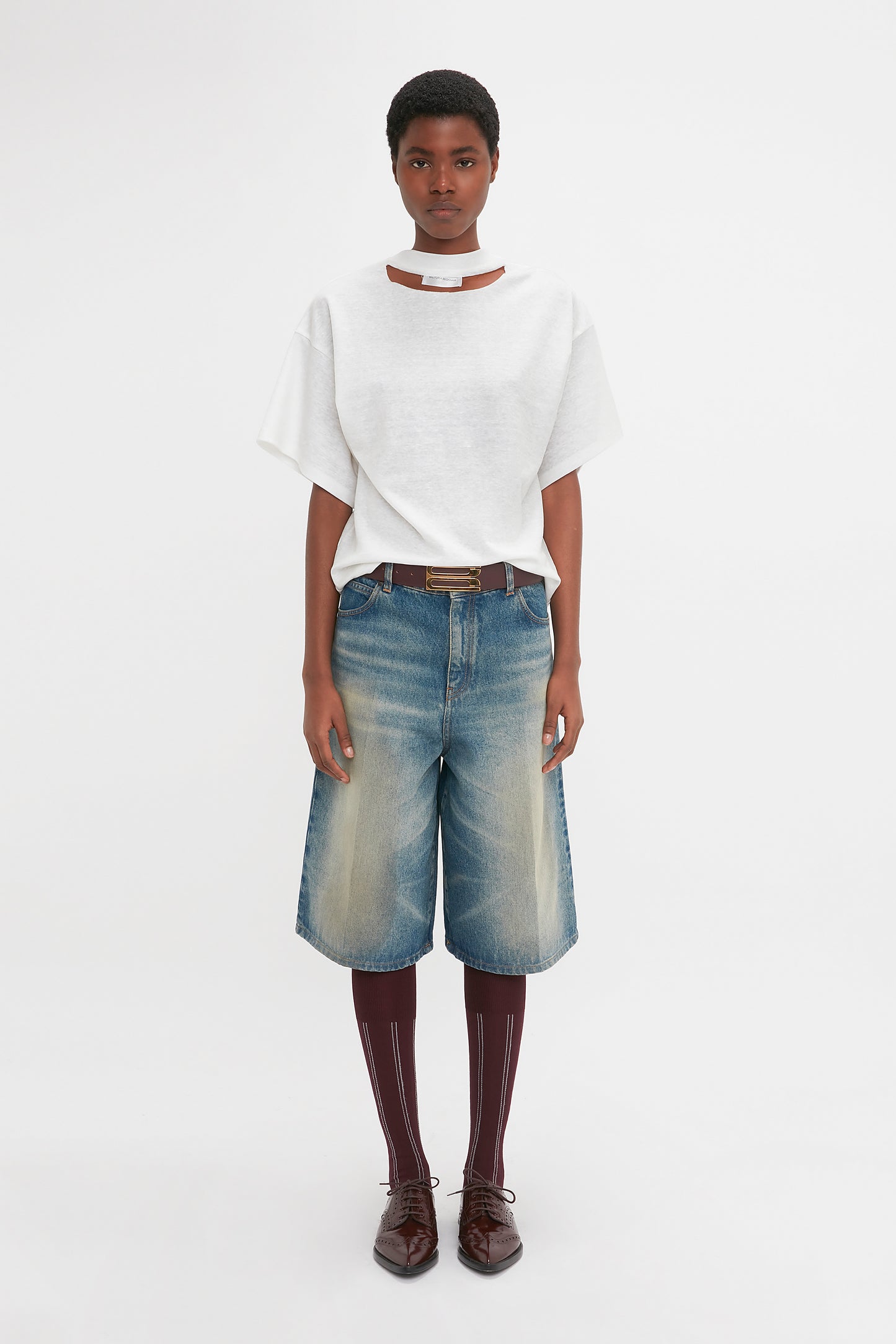 A person stands against a white background, wearing a white t-shirt and Victoria Beckham Oversized Bermuda Short In Antique Indigo Wash. They complete the look with dark socks, brown shoes, and a white choker-style necklace.