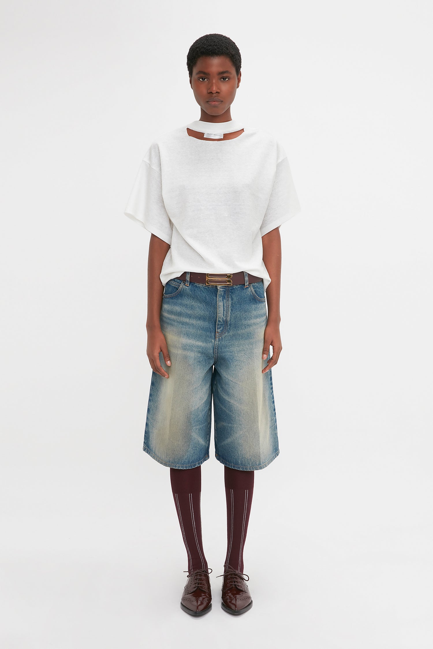 A person stands against a white background, wearing a white t-shirt and Victoria Beckham Oversized Bermuda Short In Antique Indigo Wash. They complete the look with dark socks, brown shoes, and a white choker-style necklace.