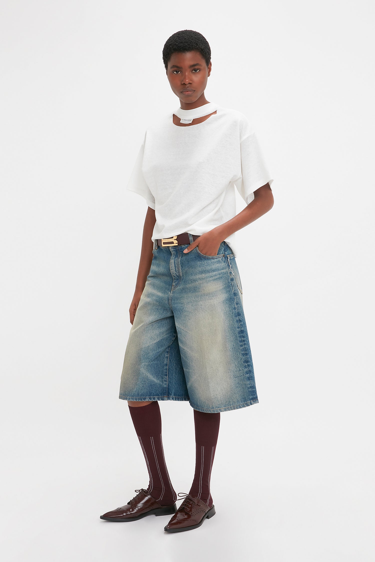 Person wearing a white T-shirt, Victoria Beckham Oversized Bermuda Short In Antique Indigo Wash, brown knee-high socks, and brown shoes with laces, standing against a plain white background.