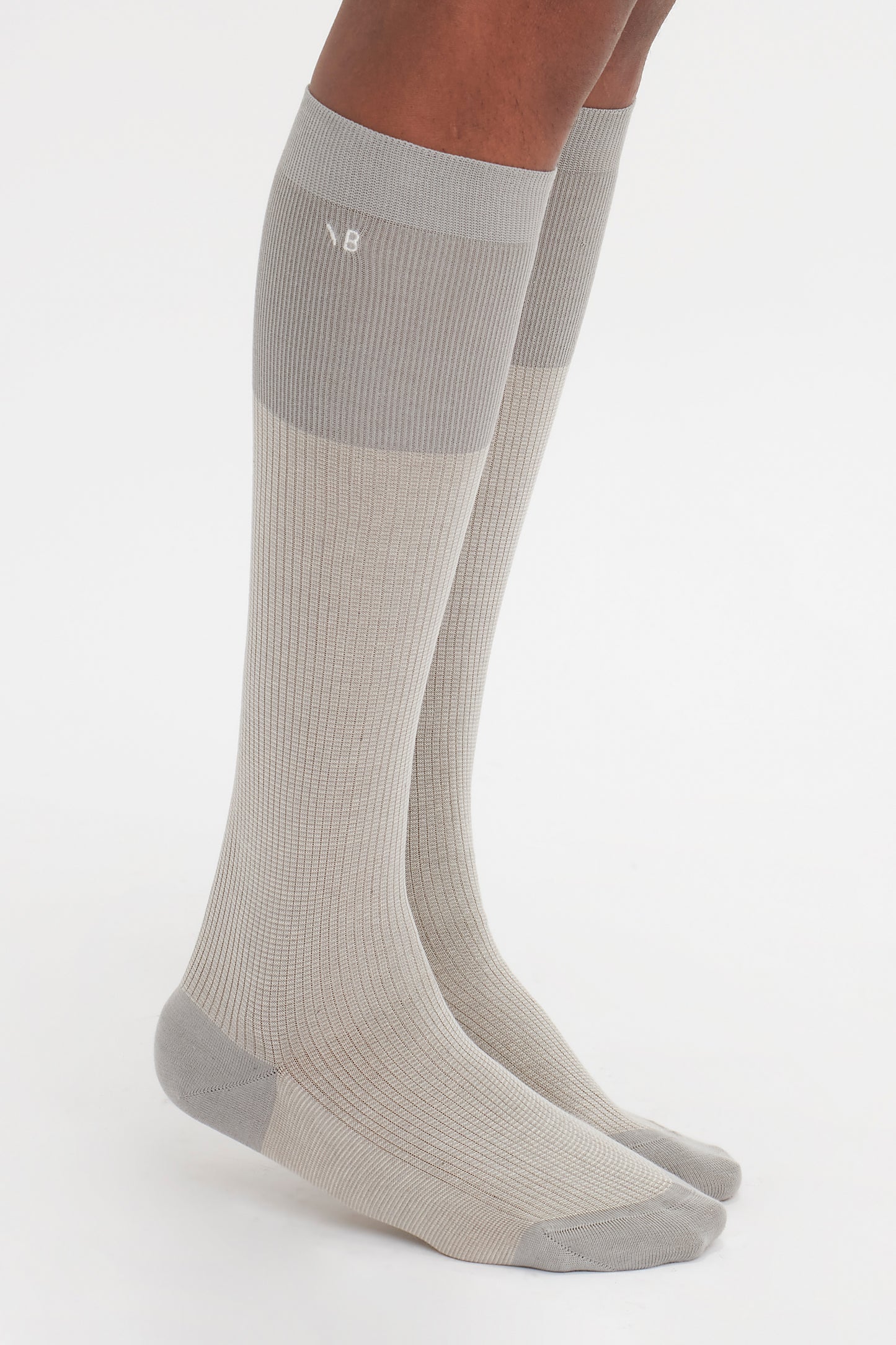 Close-up of a person's lower legs wearing knee-high, light gray superfine ribbed compression socks. The socks have a subtle two-tone design with a darker gray at the top and a white logo near the knee, crafted from 100% cotton for an elevated wardrobe essential. These are the Superfine Rib Socks In Lunar Grey by Victoria Beckham.
