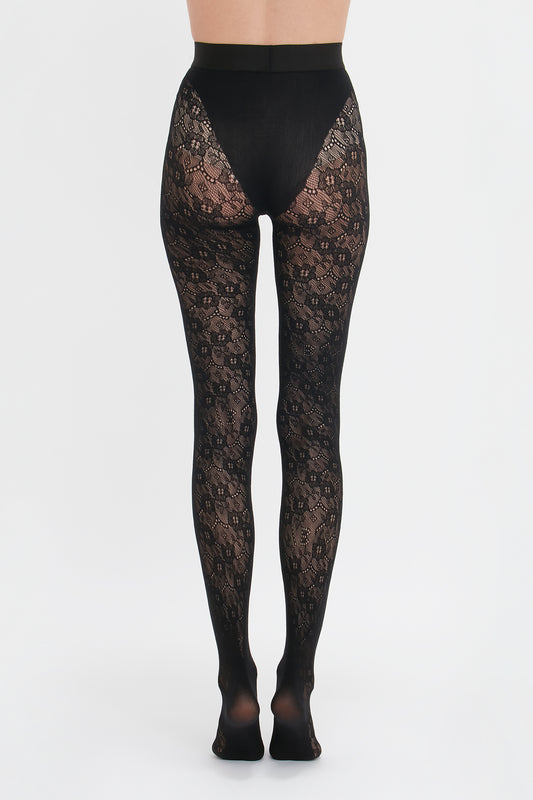 A person wearing black, intricate lace-patterned Victoria Beckham Monogram Lace Tights against a white background.