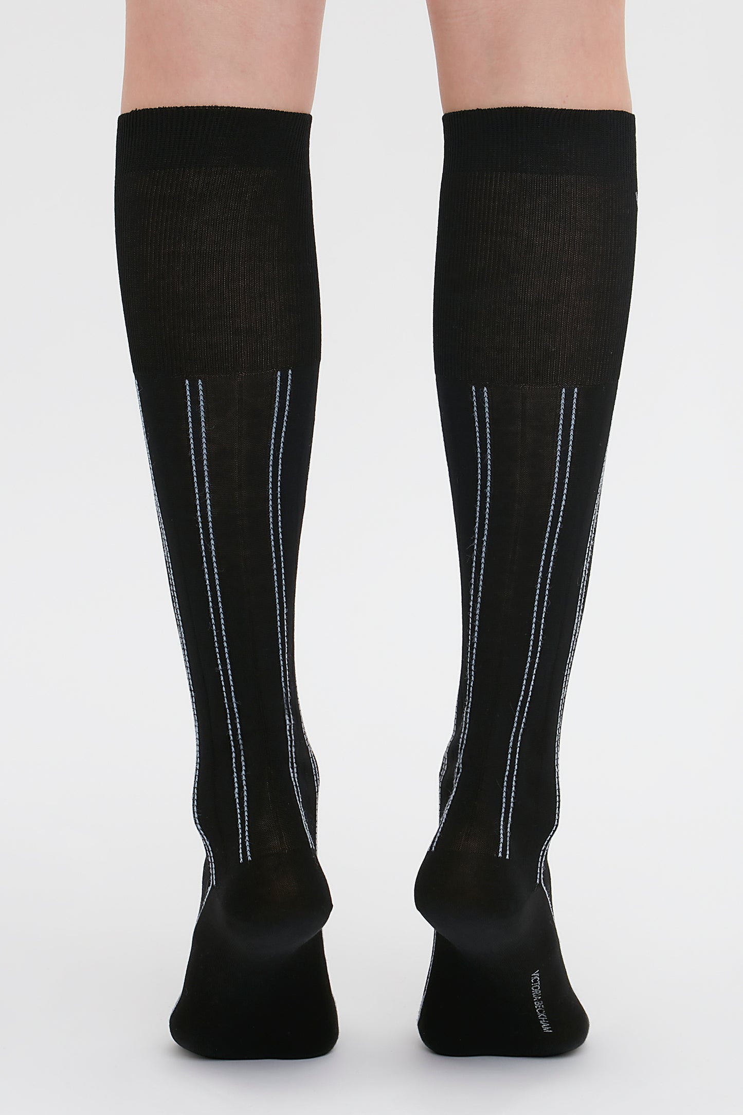 A person wearing black Superfine Rib Socks In Black by Victoria Beckham with thin blue and white vertical stripes on the back, standing on a white background.