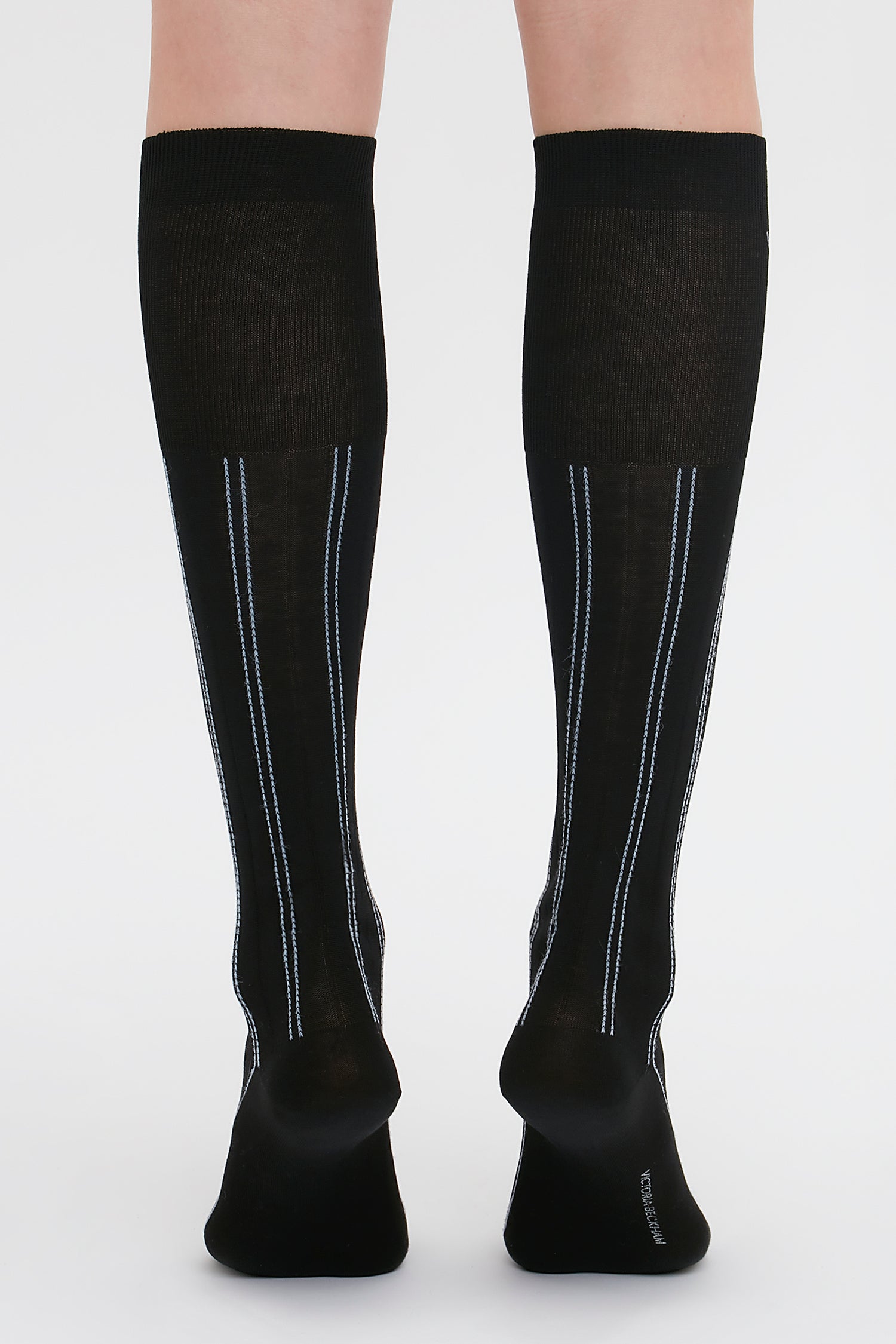 A person wearing black Superfine Rib Socks In Black by Victoria Beckham with thin blue and white vertical stripes on the back, standing on a white background.