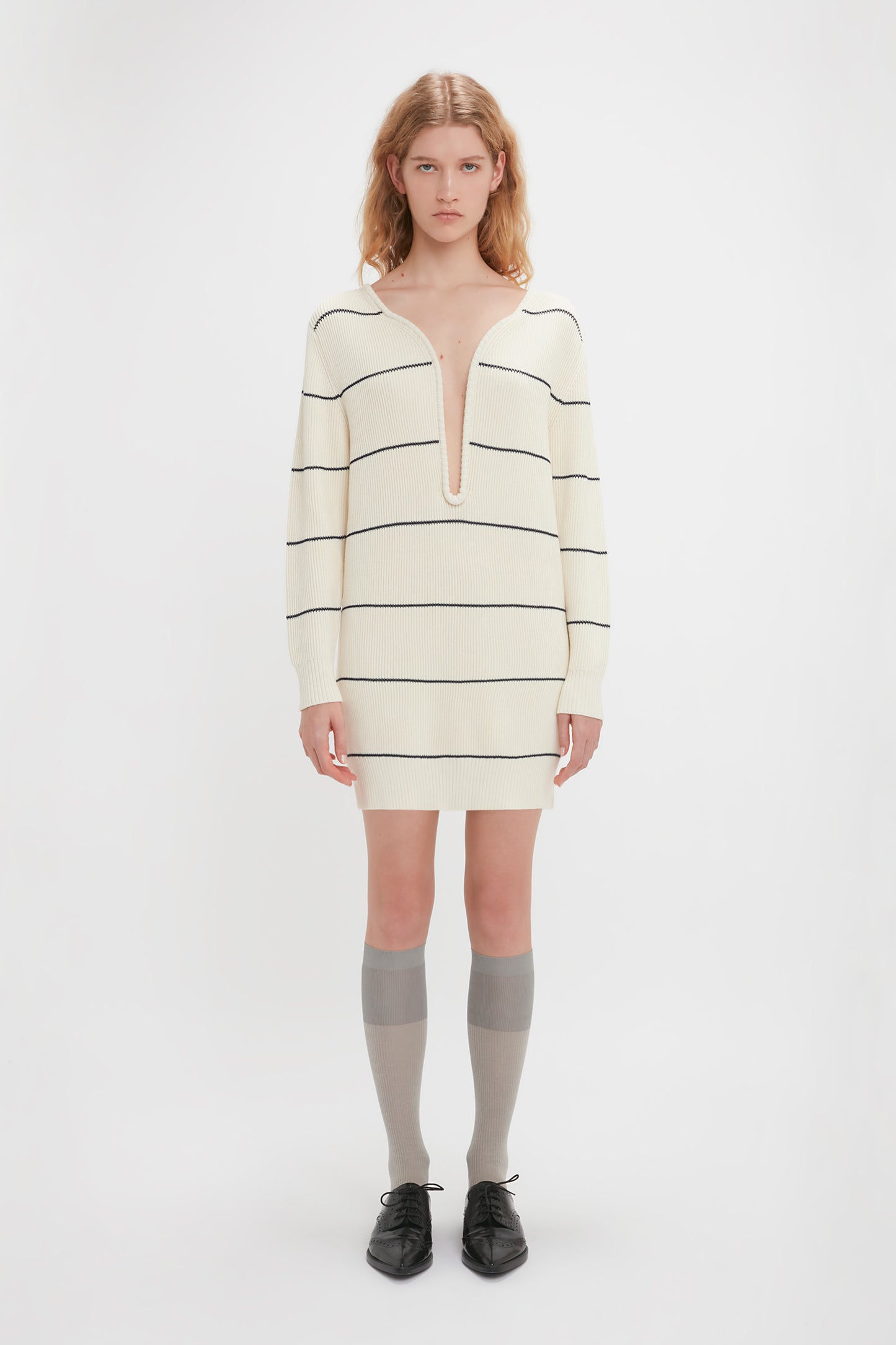 A person stands facing forward, wearing a Victoria Beckham Frame Detail Jumper Dress In Natural-Navy with black horizontal stripes, gray knee-high socks, and black shoes, against a plain white background. The mid-weight knit fabric adds texture to the attire.