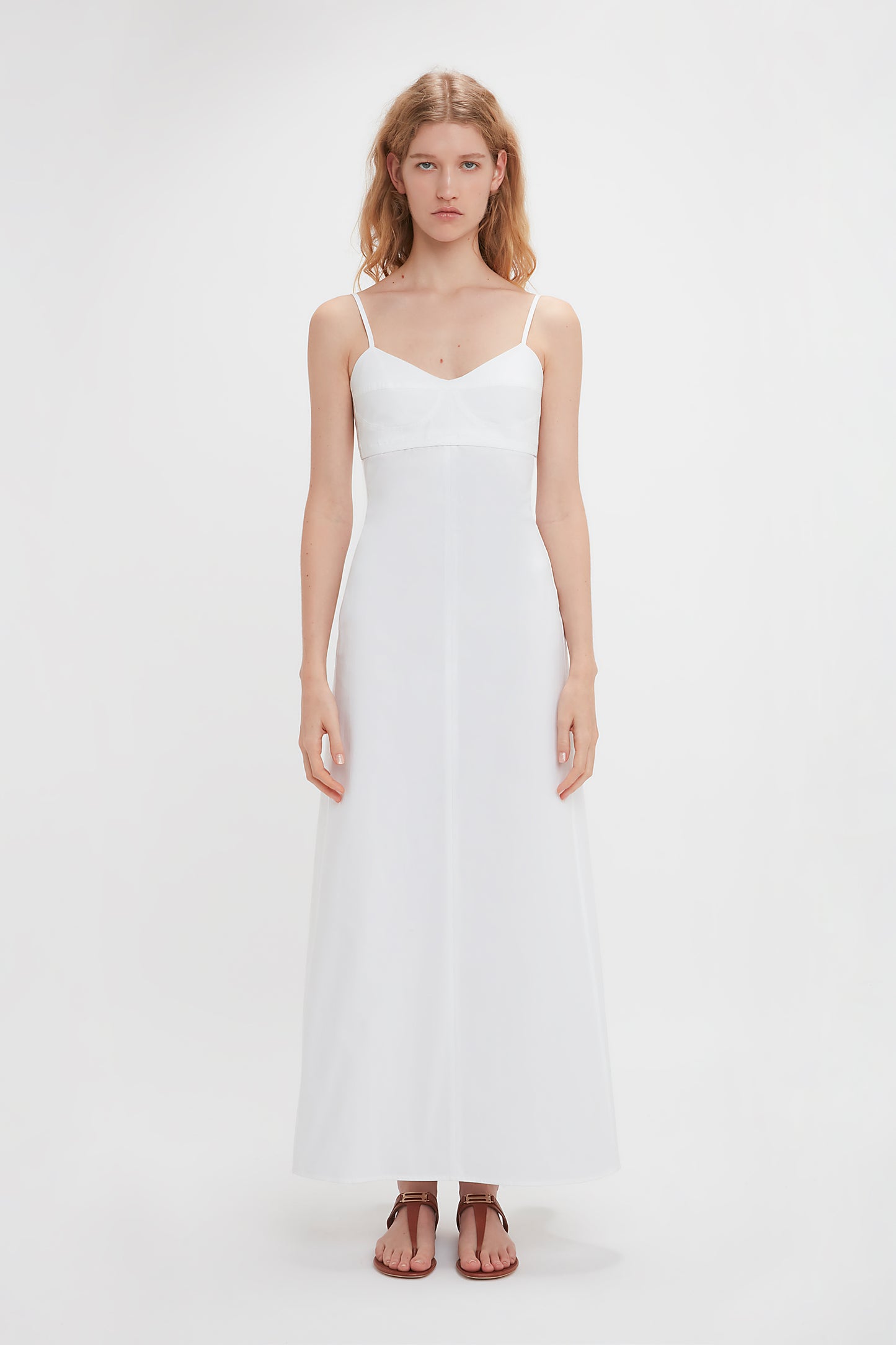 Woman in a white sleeveless dress with spaghetti straps and Flat Chain Sandal In Tan Leather by Victoria Beckham, standing against a plain white background.