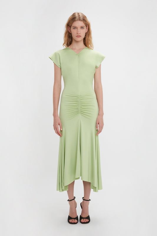 A woman in a Victoria Beckham pale green, sleeveless ruched jersey dress with an asymmetric hemline, standing against a white background, wearing black strappy heels.