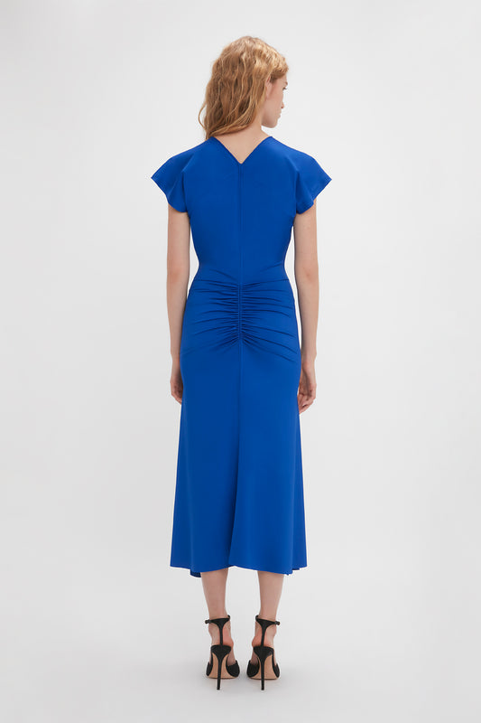 Woman standing with her back to the camera, wearing a Victoria Beckham Sleeveless Rouched Jersey Dress in Royal Blue with an asymmetric midi-length hemline and black high heels on a white background.