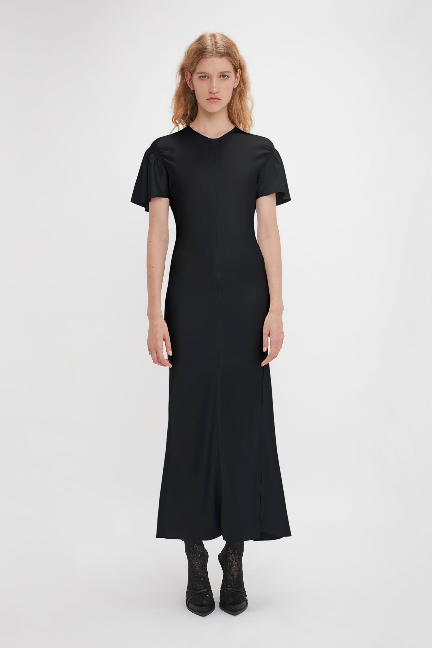 A person with long hair stands facing the camera, wearing a Victoria Beckham Gathered Sleeve Midi Dress In Black and black boots, against a plain white background.