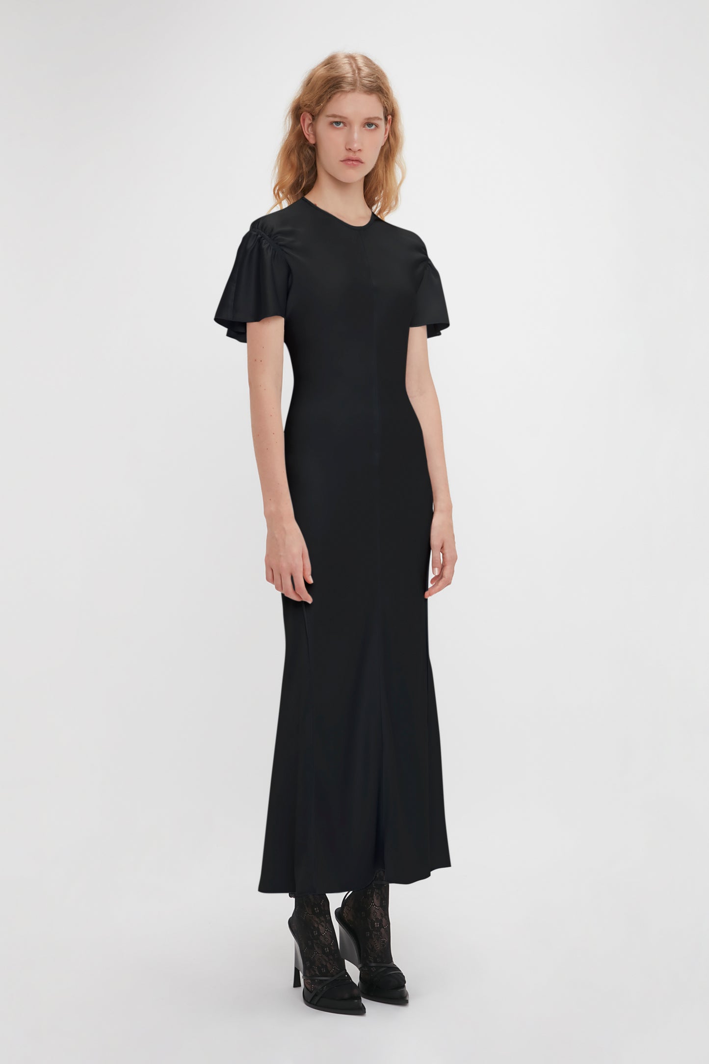A woman with long hair stands against a plain background, wearing a black Gathered Sleeve Midi Dress In Black by Victoria Beckham and black heeled shoes. The crepe back satin fabric of the dress adds a subtle sheen. She is facing the camera with a neutral expression.