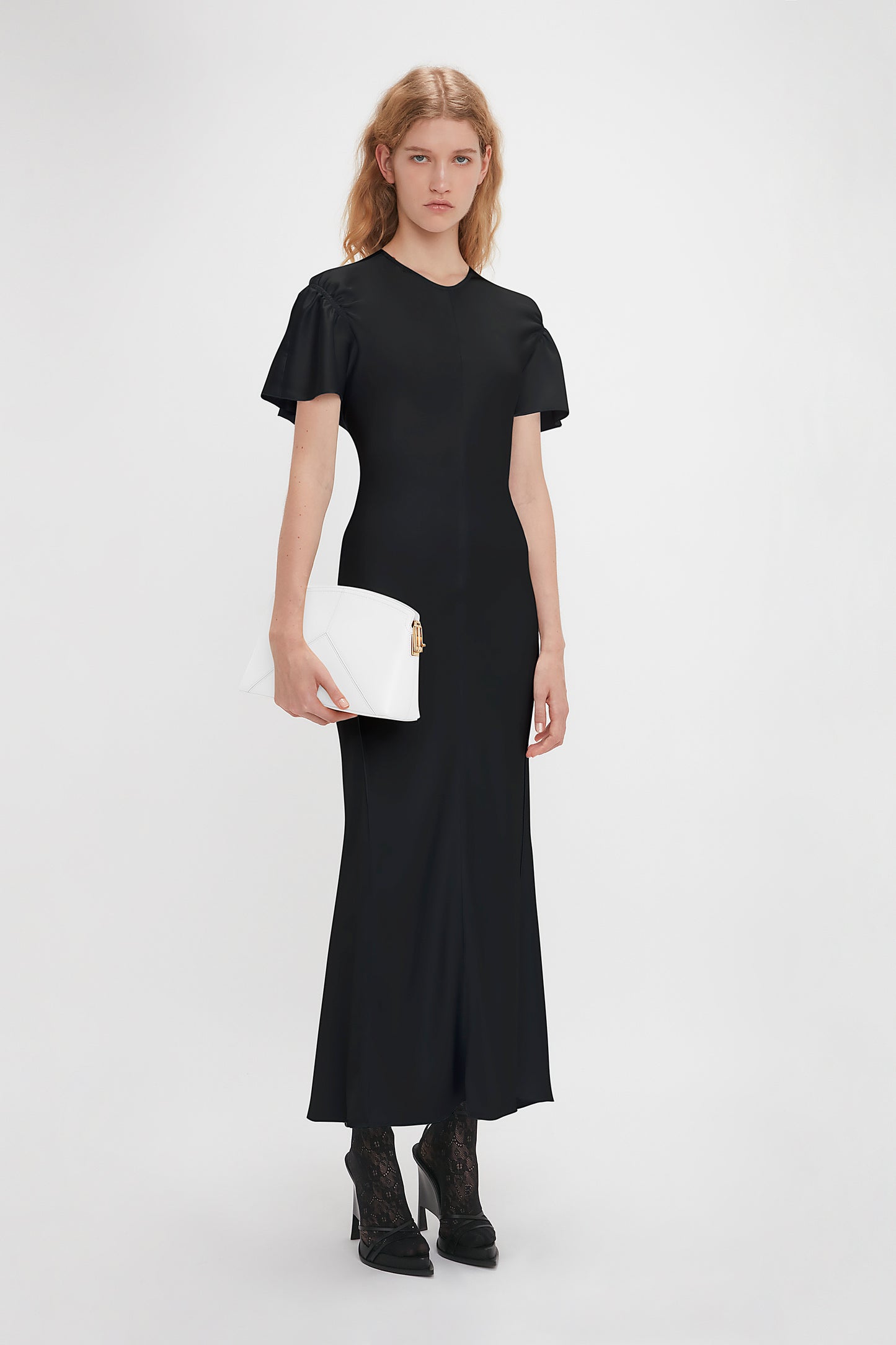A person wearing a Gathered Sleeve Midi Dress In Black by Victoria Beckham and black ankle boots holds a white clutch against a plain white background.